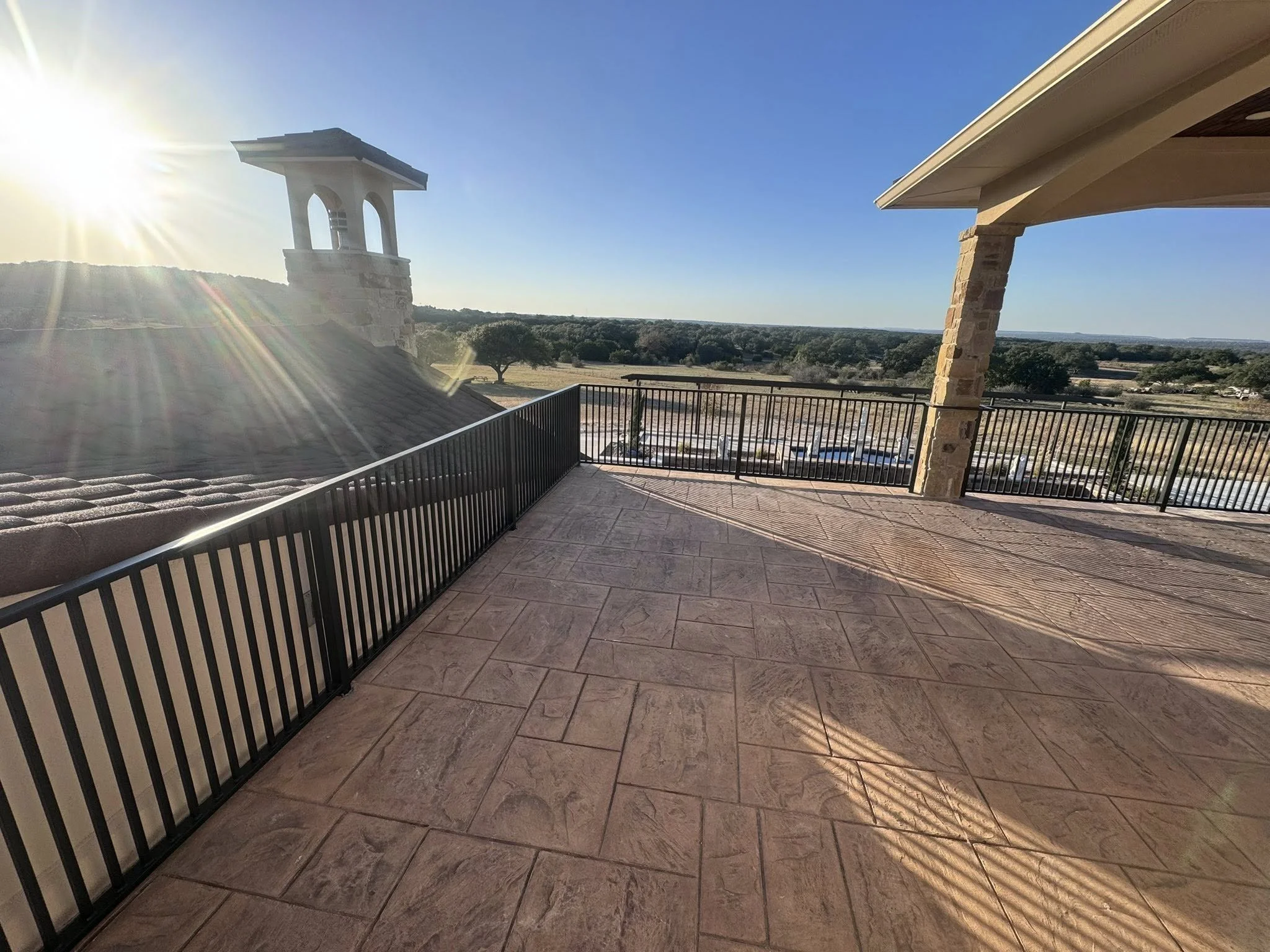 Balcony with tiled floor, black metal railing, stone columns, overlooking a rural landscape with trees and open fields, during sunset.