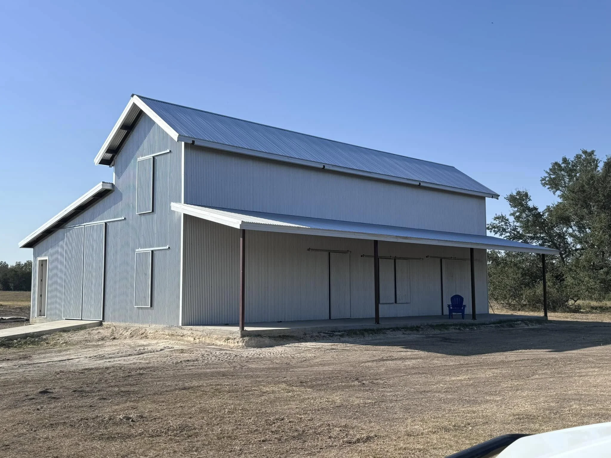 A large, metal barn with a blue roof and vertical siding, situated on a dirt lot with a few trees in the background and a blue chair on the porch.