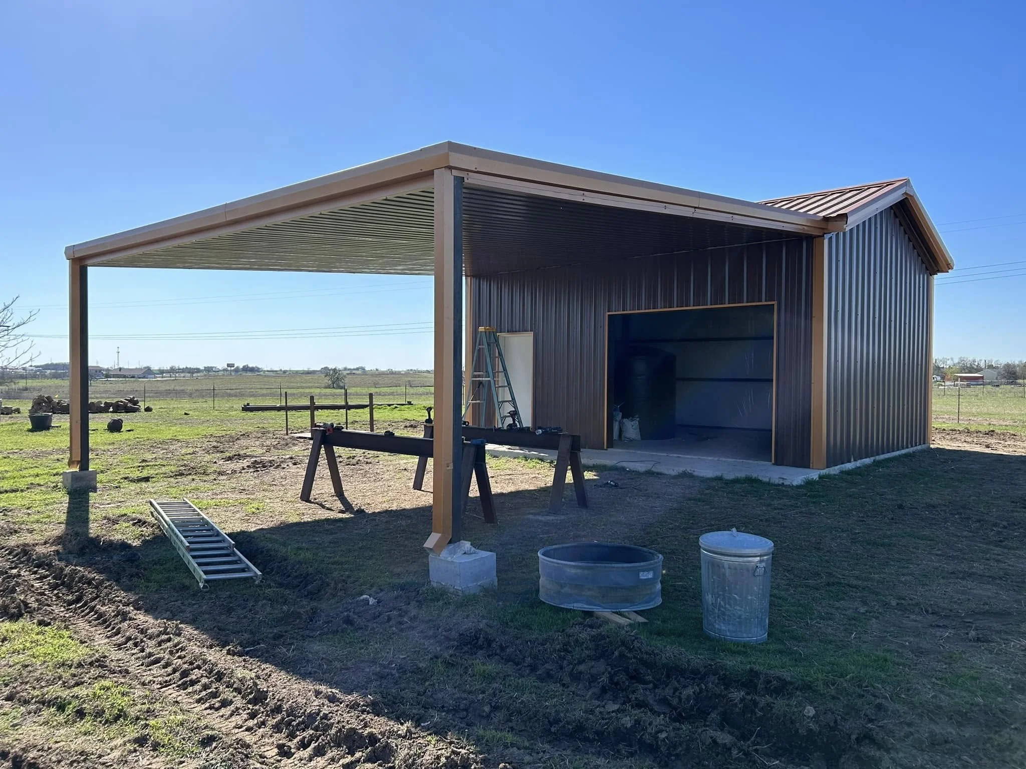 A partially built metal shed on a farm with a raised roof extension supported by metal post, a ladder leaning against the shed, and construction tools and materials on the ground.