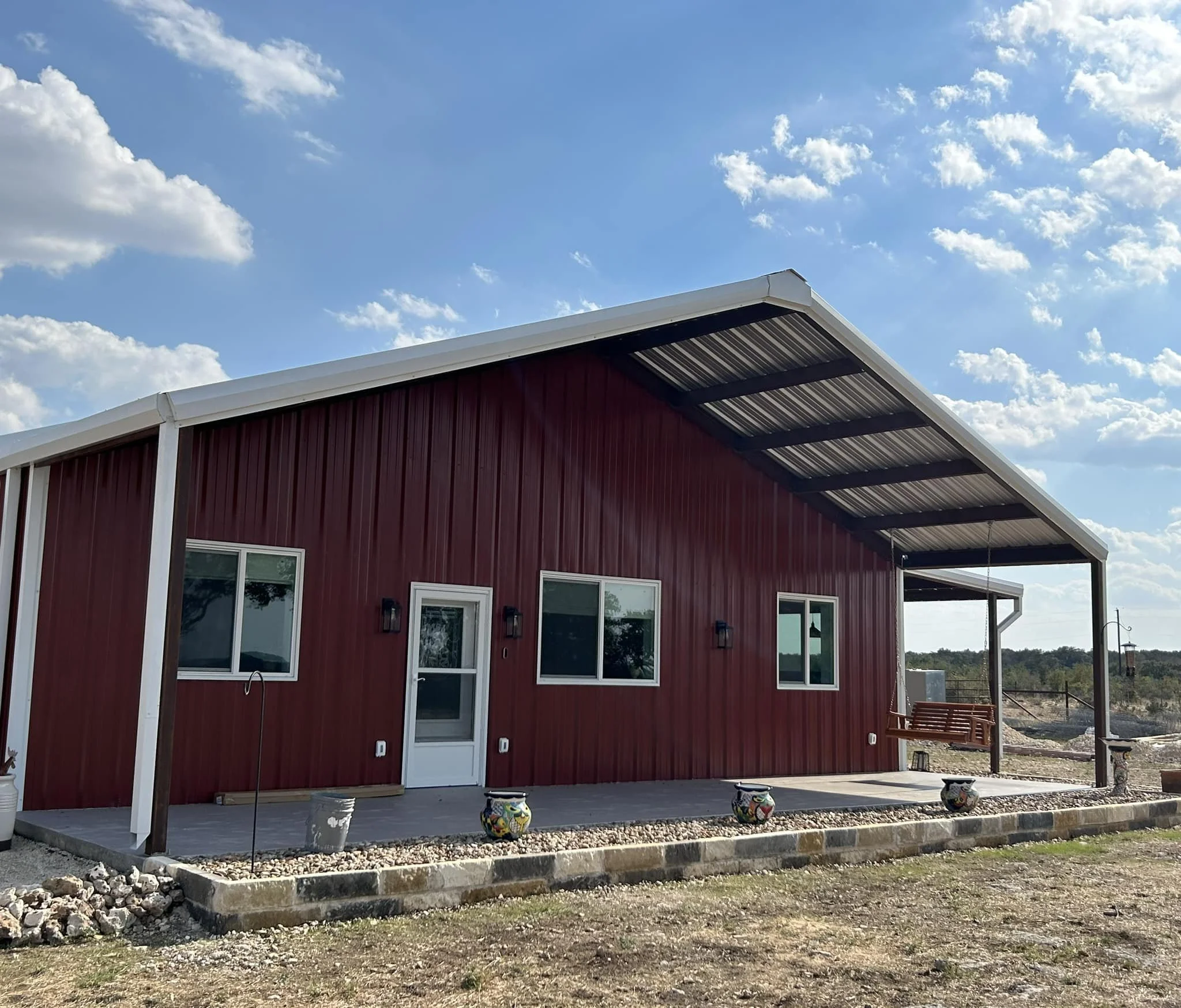 A red metal house with a sloped roof and a covered porch, sitting on a concrete slab with potted plants, under a partly cloudy sky.
