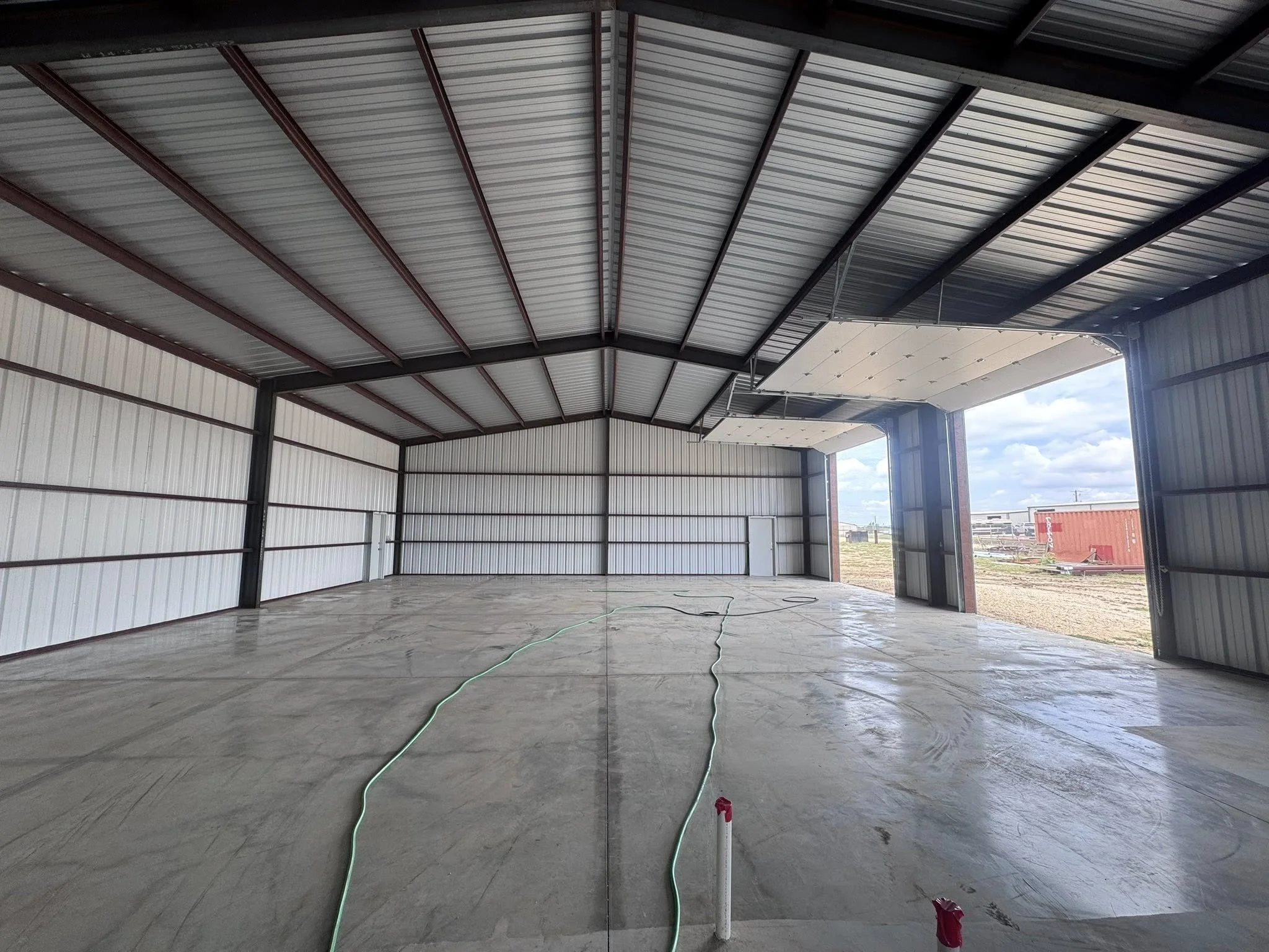 Interior of a new metal storage building or warehouse with concrete floor, partially open garage doors, and cityscape visible outside on a partly cloudy day.