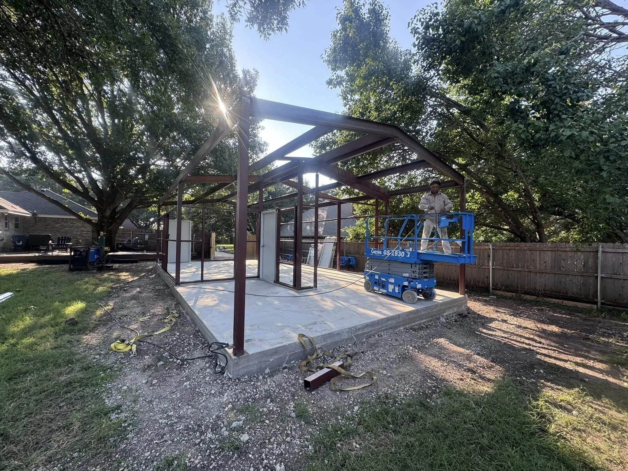 A construction worker stands on a blue lift in a backyard, working on a small building's metal frame structure with a concrete foundation, surrounded by trees and a wooden fence.