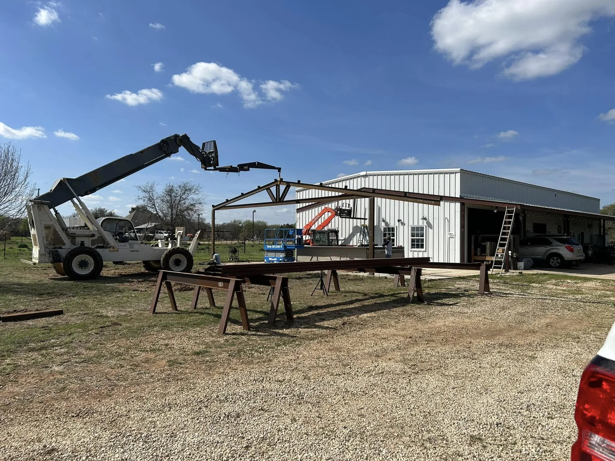 Building a metal framework outside a white warehouse under a blue sky with scattered clouds, with construction equipment and a wooden sawhorse in the foreground.