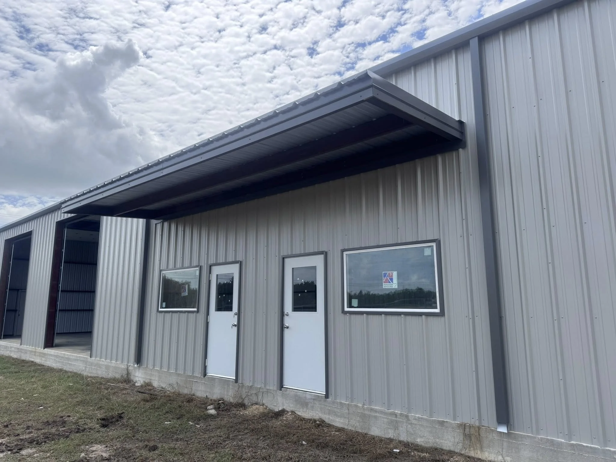 Metal building with silver siding, three windows, two doors, and a small overhang on the roof under partly cloudy sky.