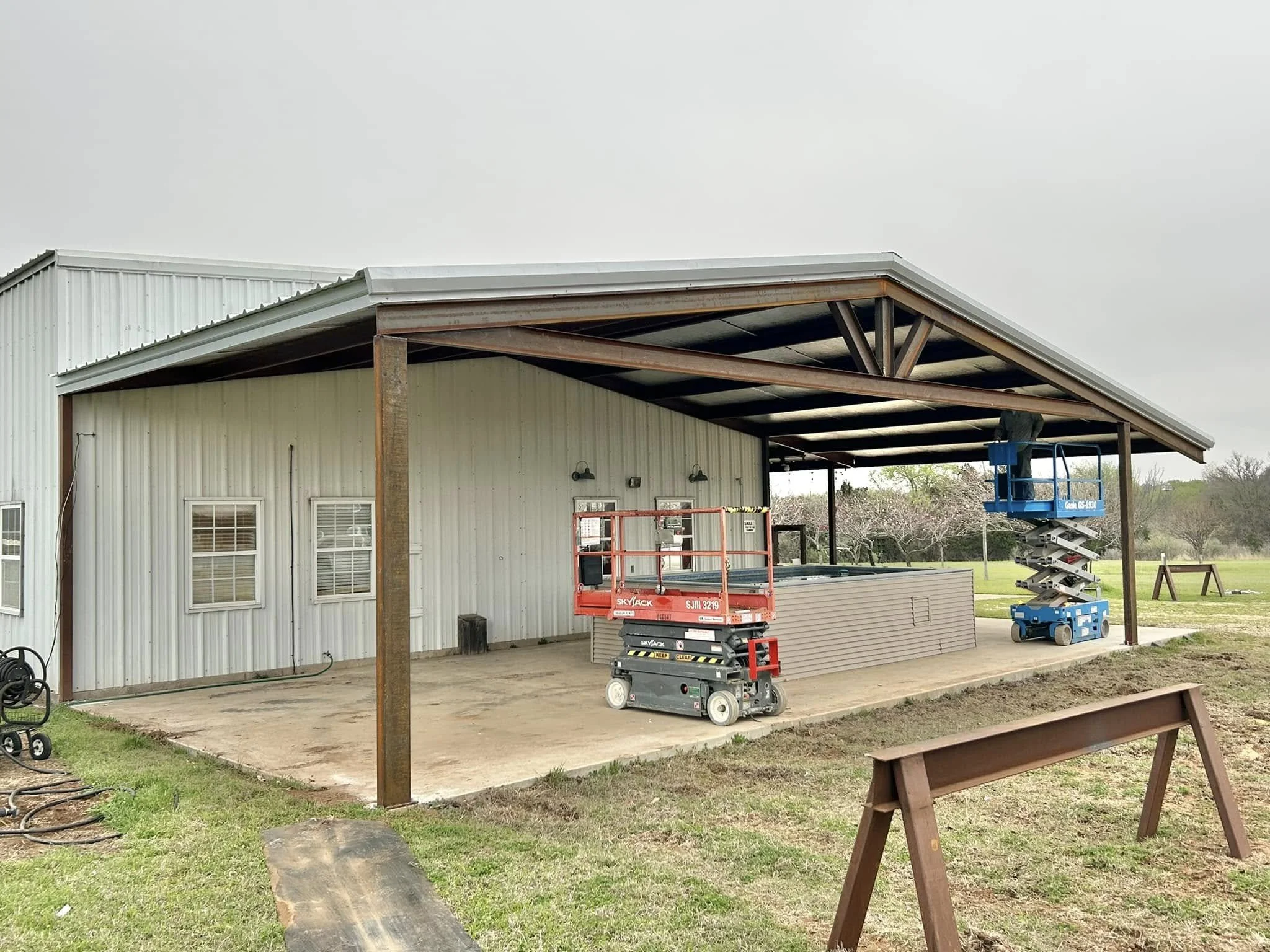 A metal building with a large covered patio area under construction. There are two scissor lift platforms, one red and one blue, positioned on the patio. The background shows a grassy area with trees and cloudy sky.