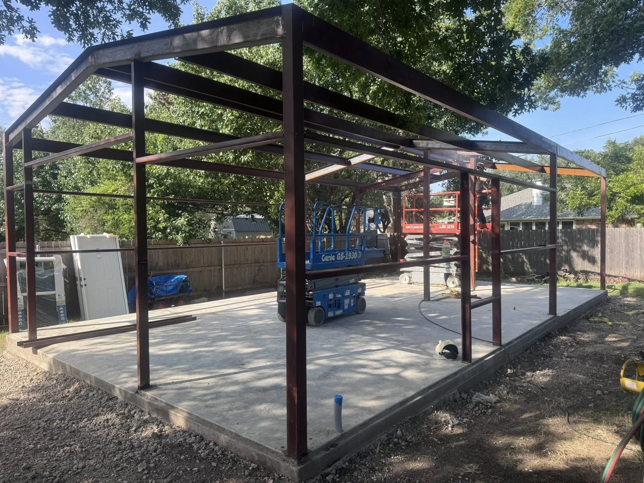 Construction site with metal framework for a building on a concrete slab, surrounded by trees and a wooden fence, with construction equipment and tools present.