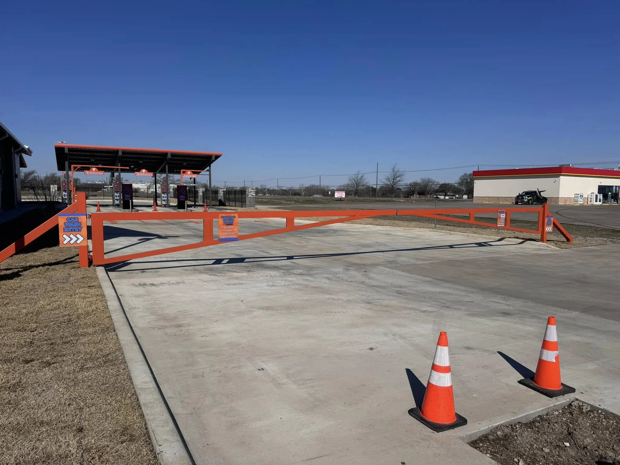 Drive-thru car wash with orange barrier gate, orange cones, and a canopy with washing stations in the background on a sunny day.