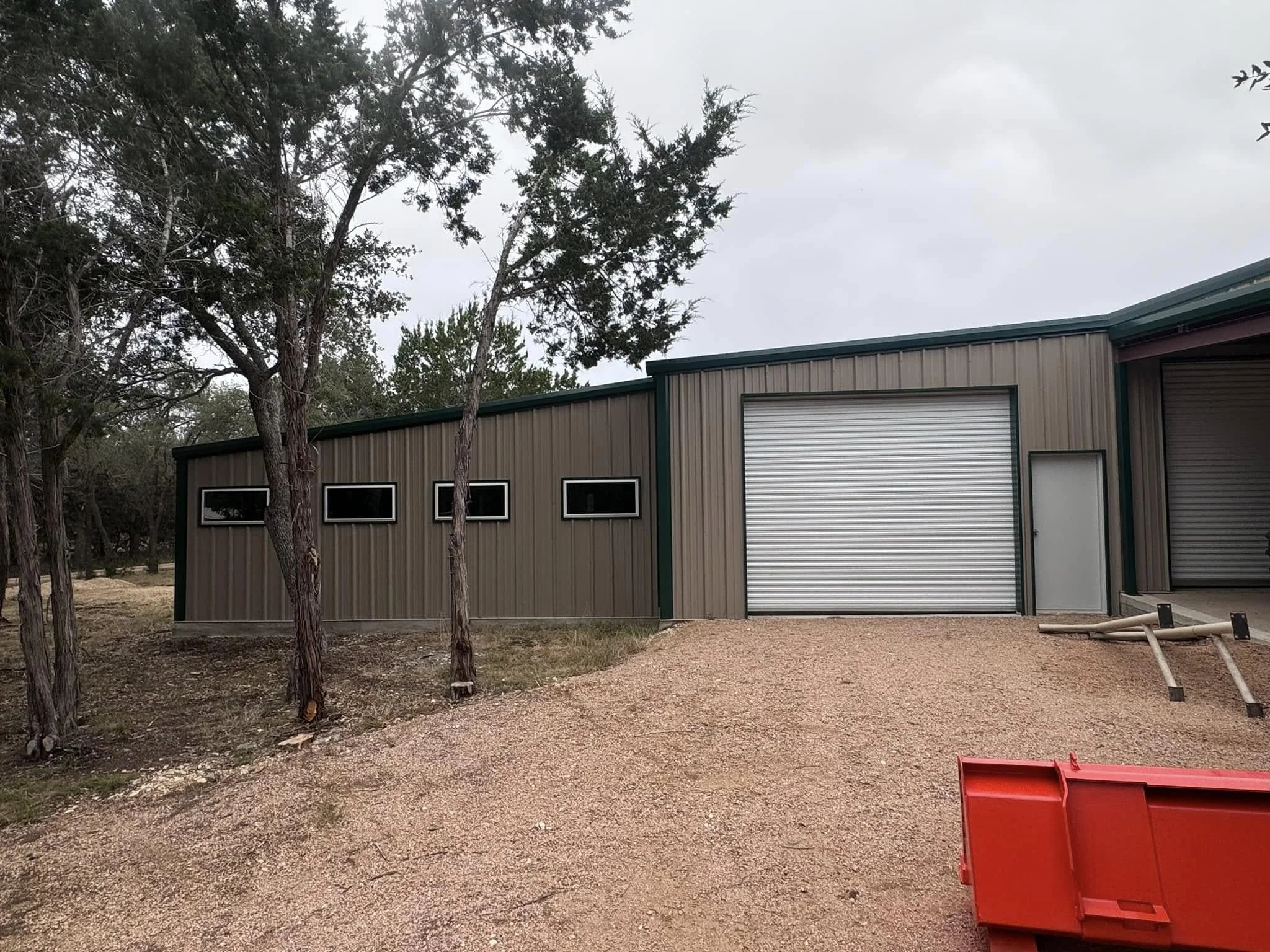 Industrial building with a large garage door, smaller door, three small horizontal windows, and trees nearby.