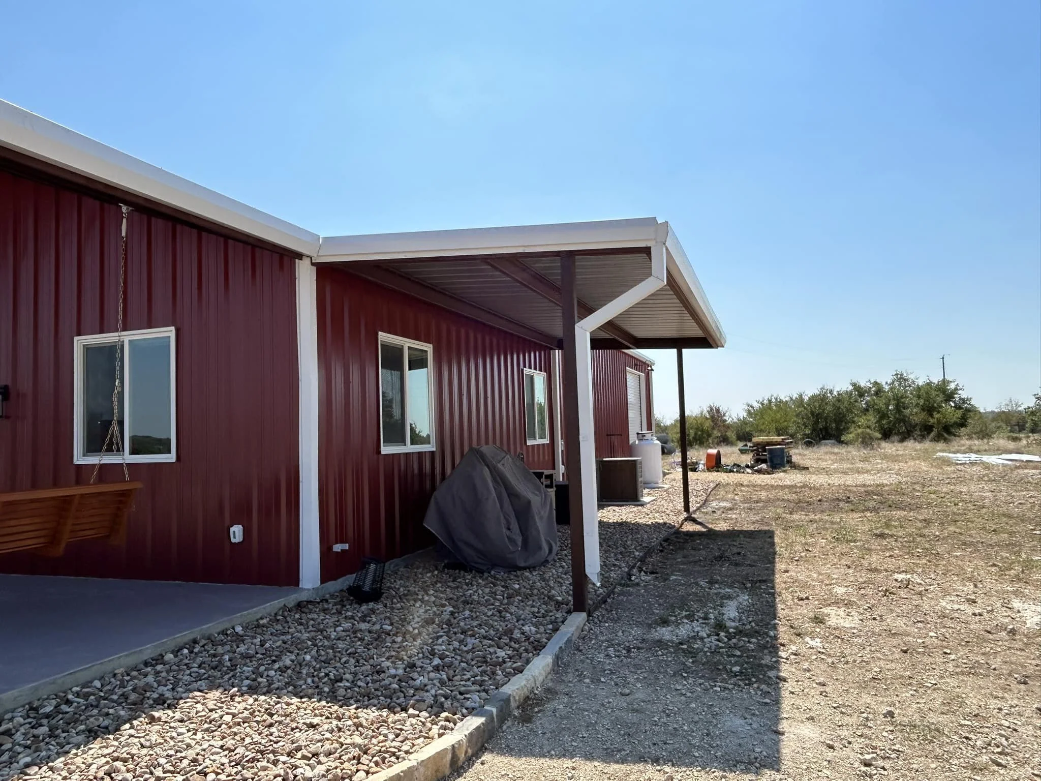 Red metal-sided house with a small porch, two white-framed windows, and a covered area supported by a post. The yard is mostly dirt and rocks with some outdoor items, and there are bushes and open land in the background under a clear blue sky.
