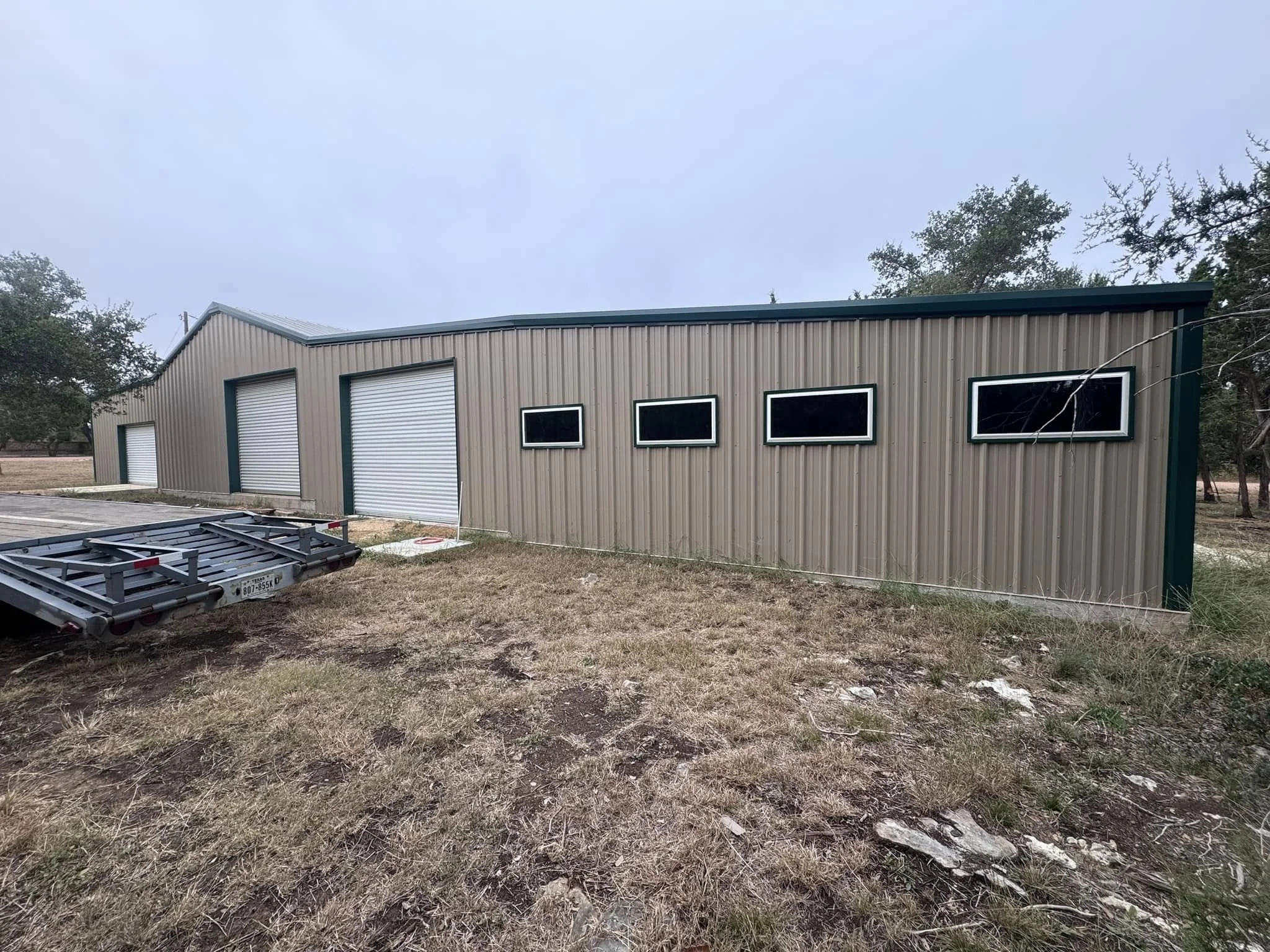 A long, tan, metal building with three large roll-up garage doors and four small, rectangular windows along its side. There is a trailer parked in front of the building on a grassy area with patches of dirt.