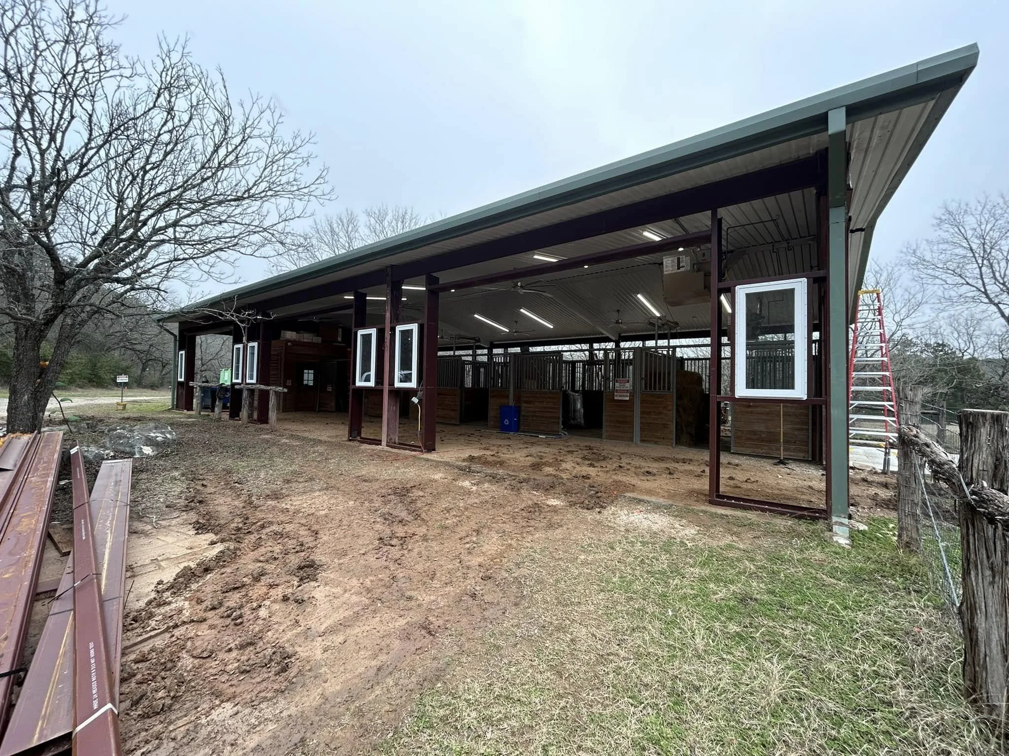 Construction of a metal barn with open sides, some windows, and a dirt ground with patches of grass, surrounded by trees.