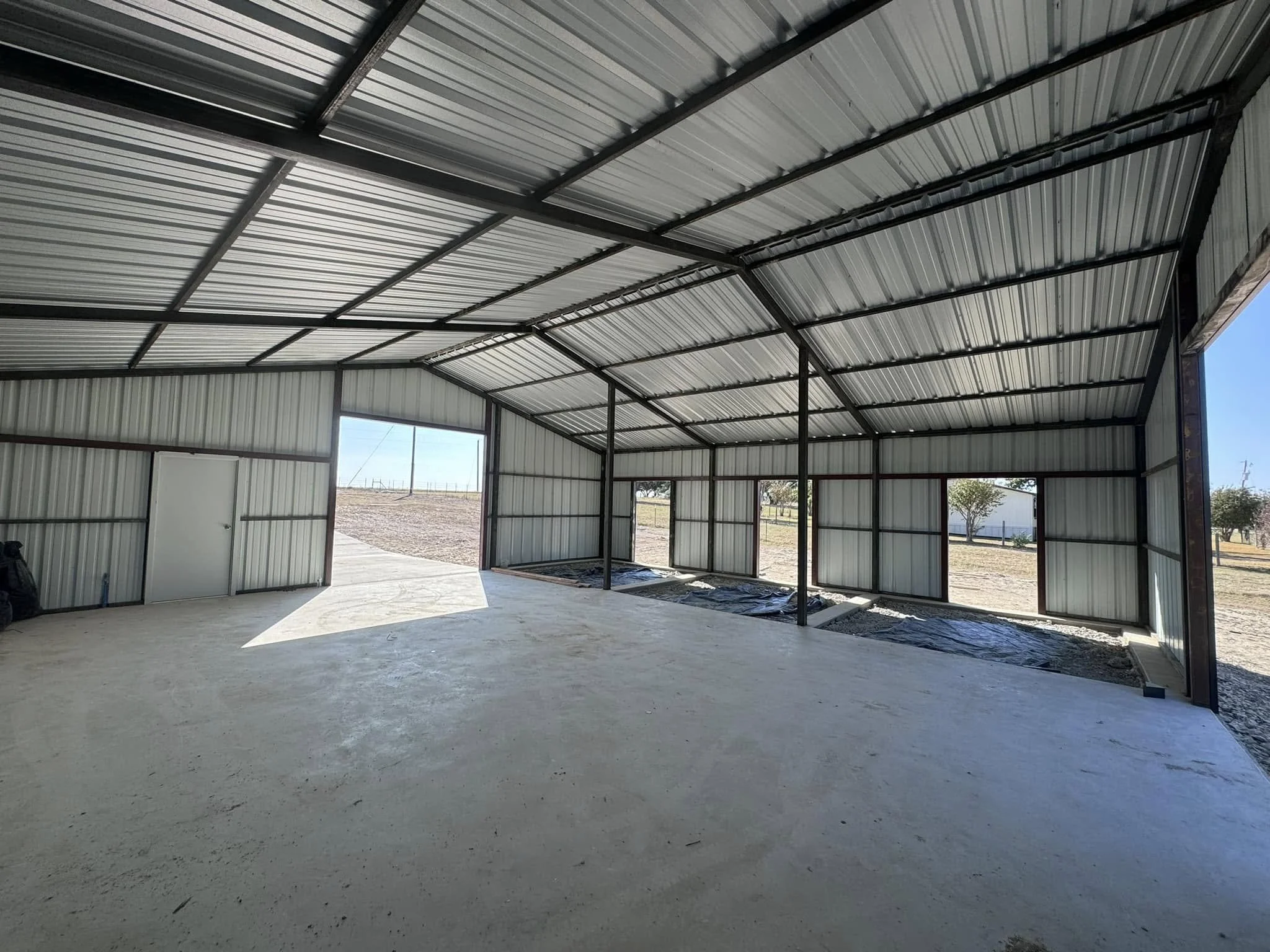 The interior of a large metal building under construction with a concrete floor, metal walls, and ceiling, showing open doorways and windows with a view of the outdoor landscape.