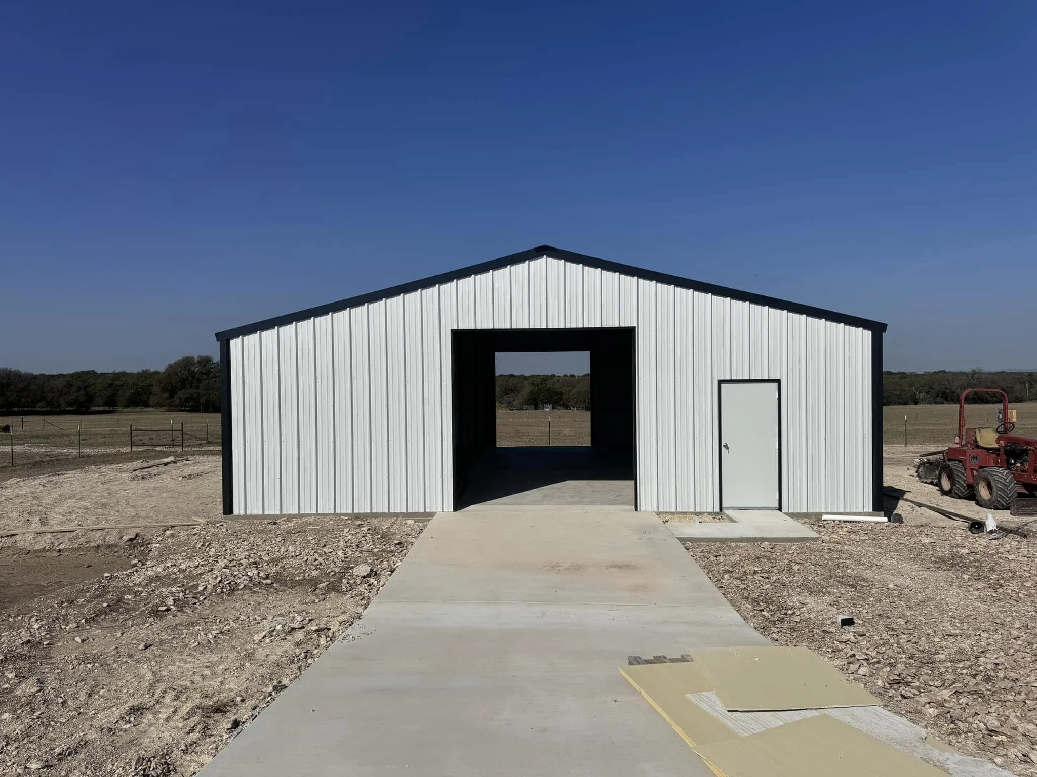 A white metal storage shed with black trim on a construction site with a concrete driveway, in a rural area with open fields and trees in the background, under a clear blue sky.