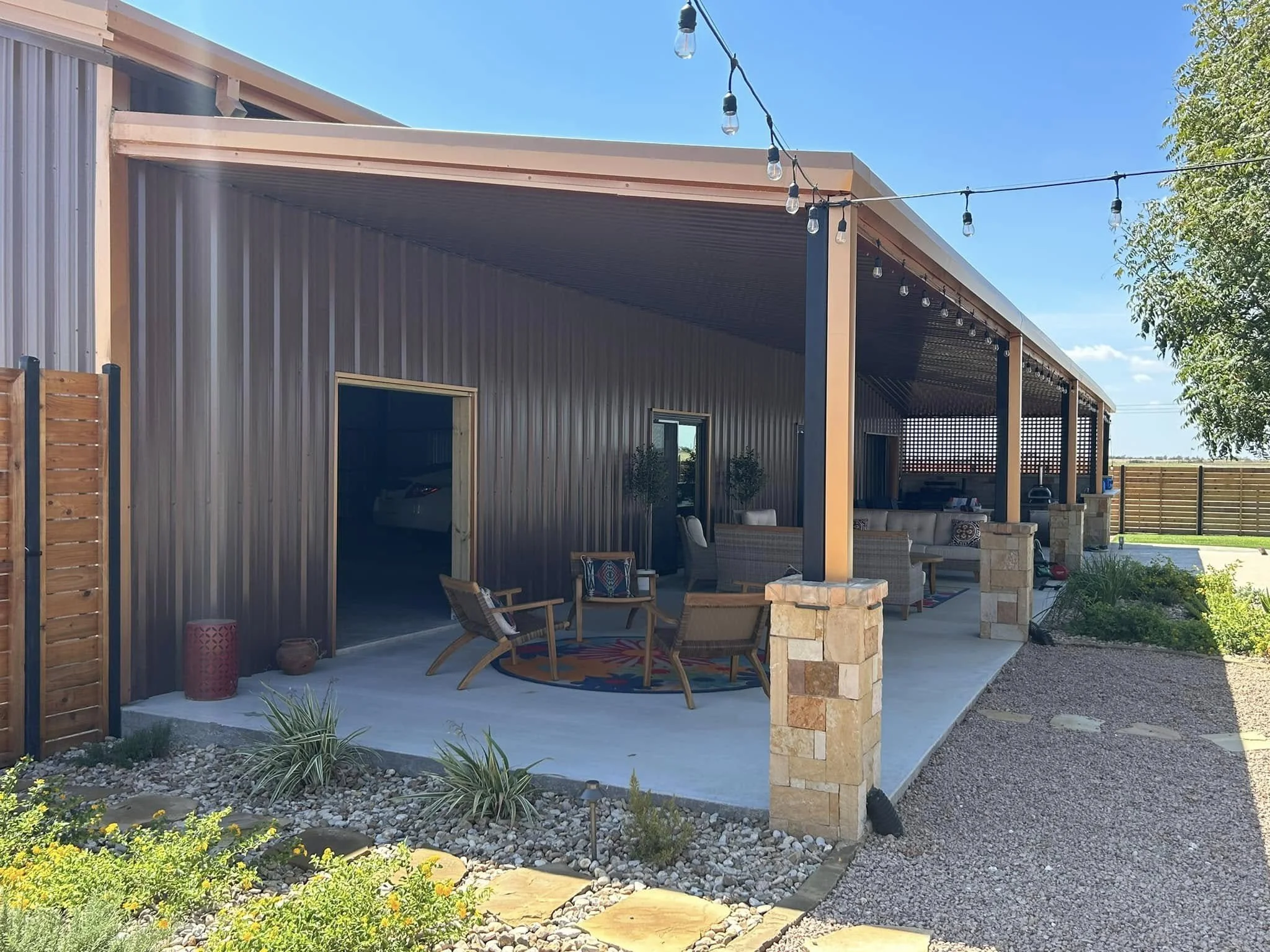 Outdoor patio with seating area, string lights overhead, and a metal building with a covered porch, surrounded by landscaped yard and gravel path.