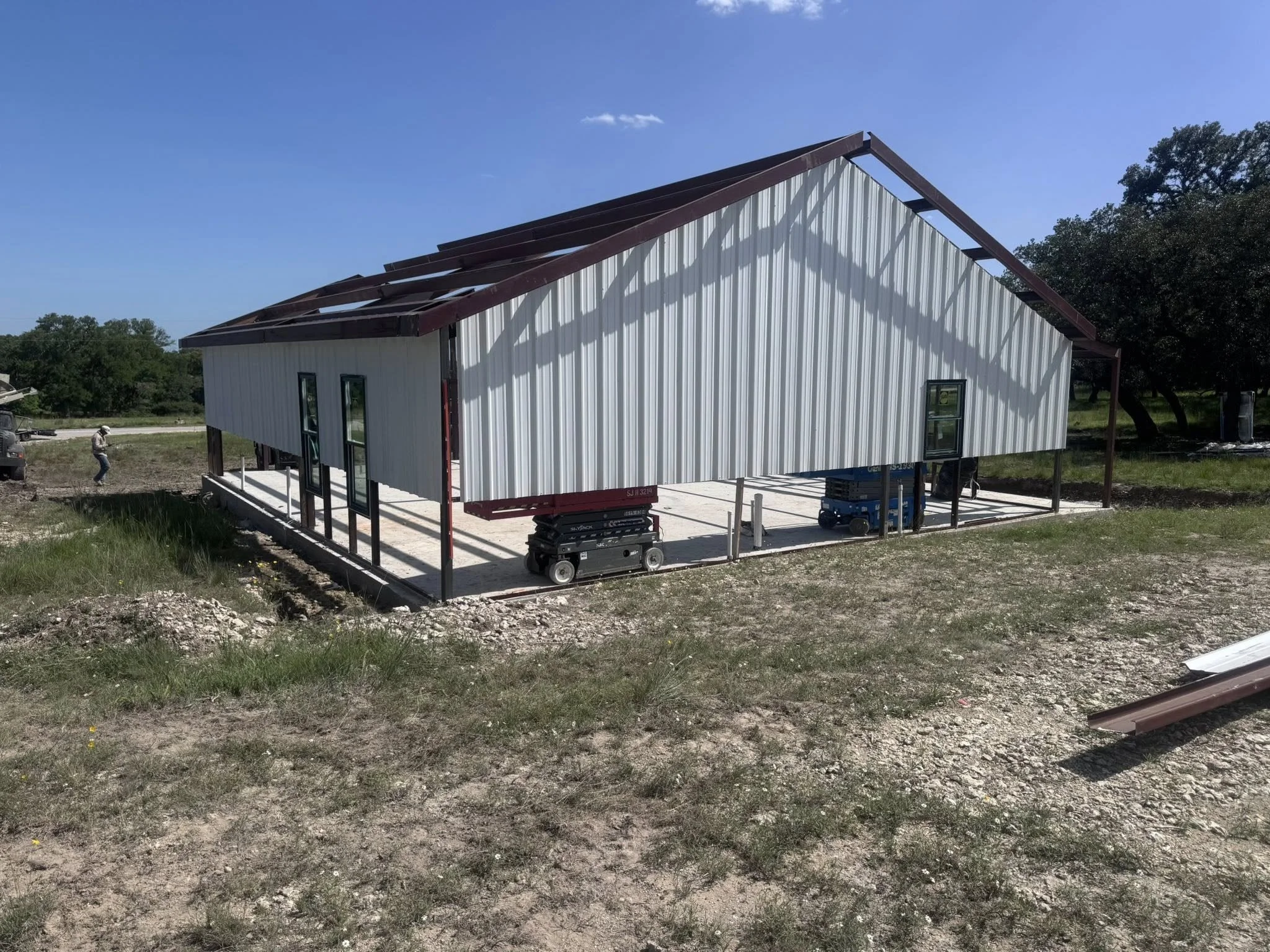A house under construction with metal siding and a sloped roof frame, situated on a dirt lot with construction equipment nearby.