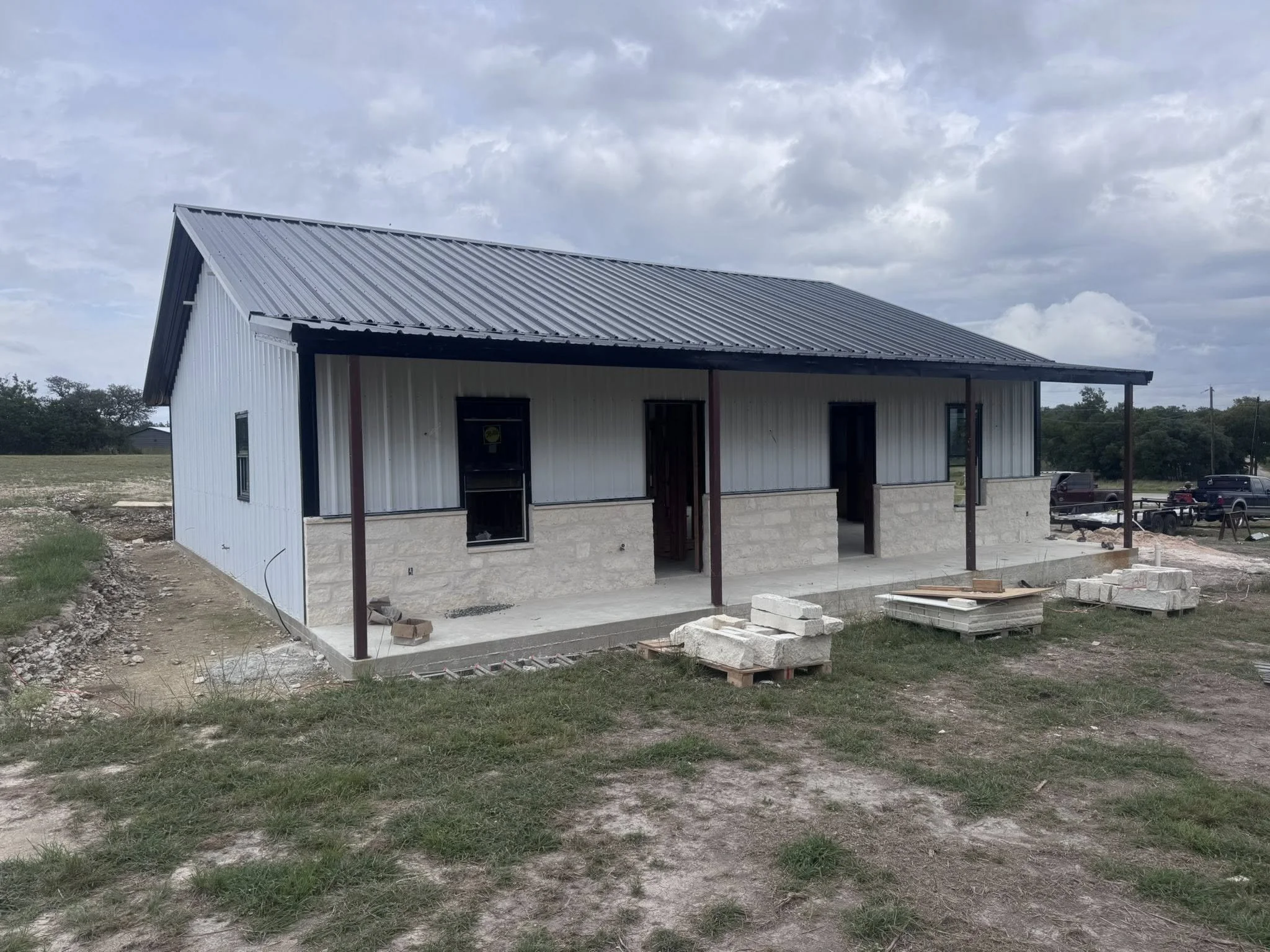 Side view of a newly constructed building with a metal roof, metal siding, and concrete block foundation. Construction materials are placed on the ground nearby. The building has a porch area supported by metal posts, with some completed sections. Th