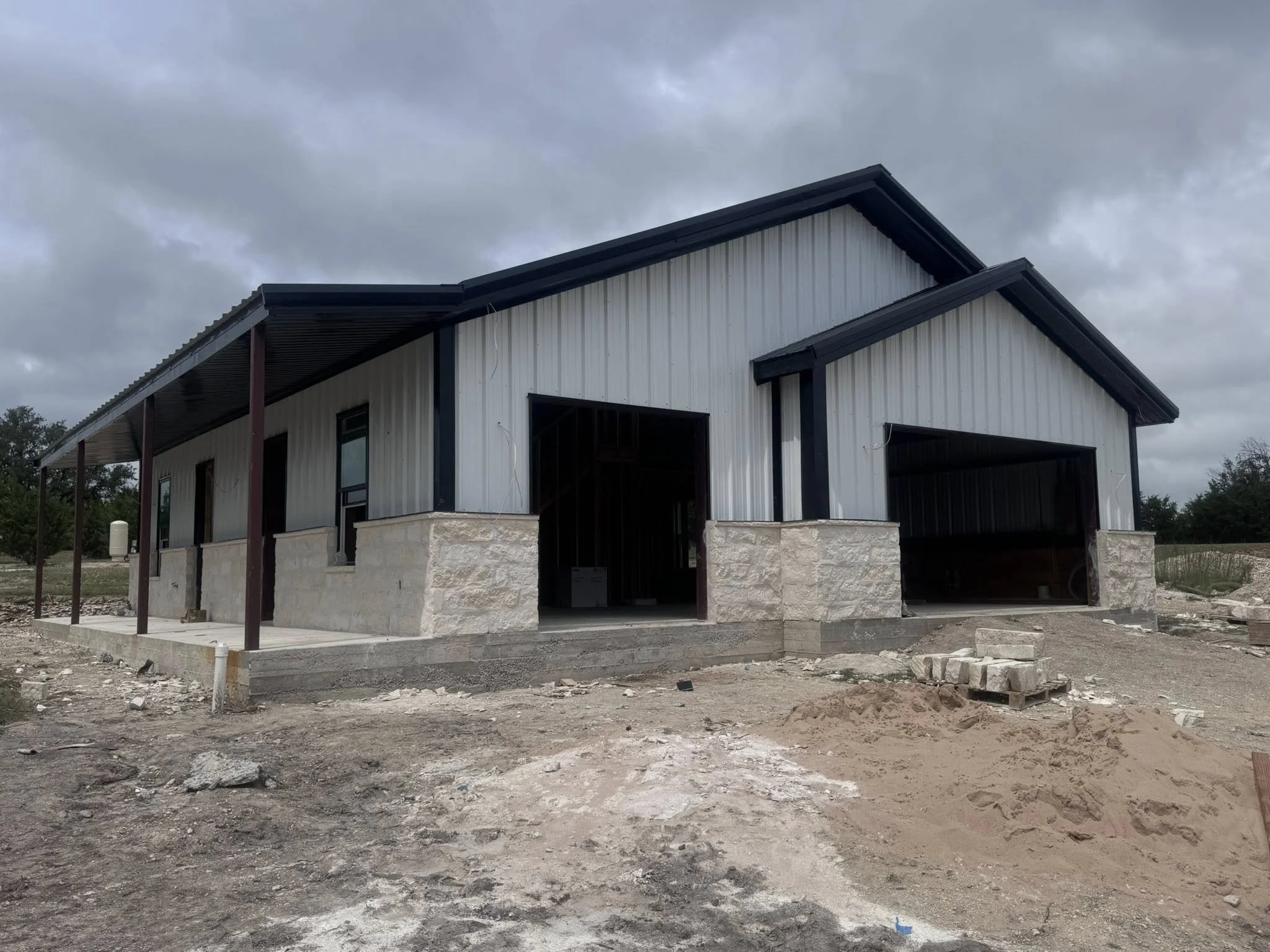 Under construction house with metal siding, stone foundation, and a carport with a metal roof. The yard is unfinished with dirt and construction materials.