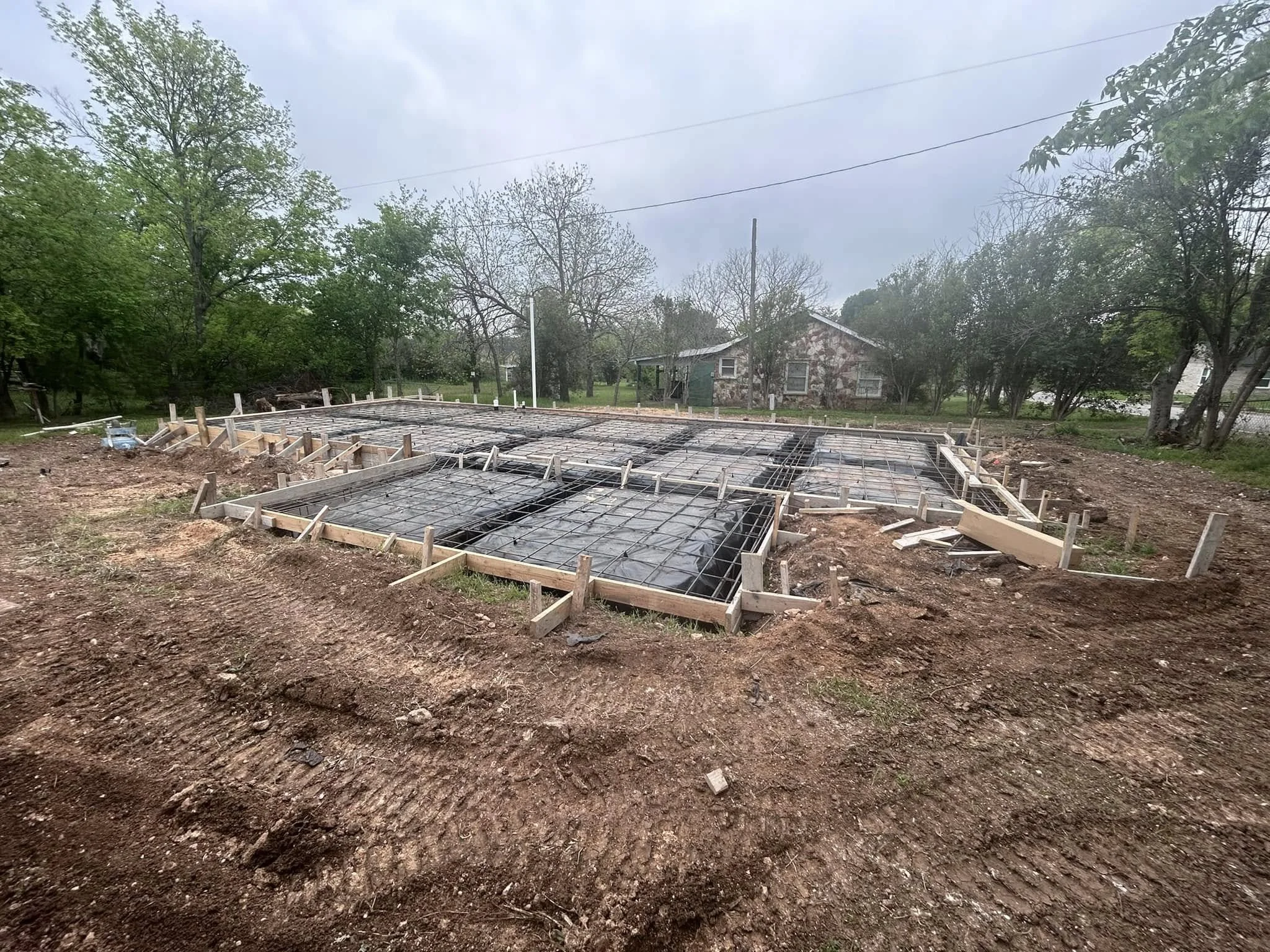 Construction site with foundation framing and rebar for a new building, surrounded by dirt and trees in the background.