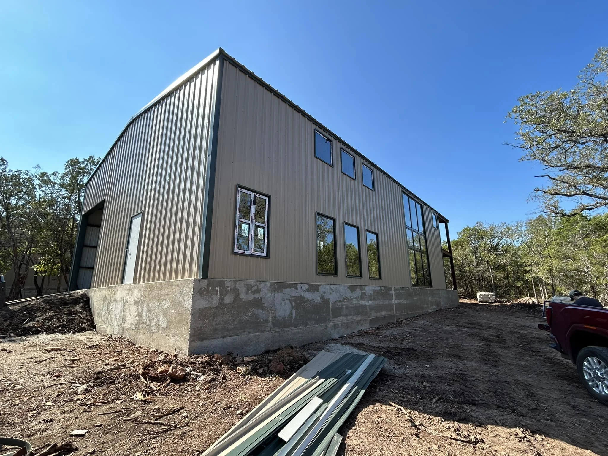 Under construction modern metal building with large windows, set on a concrete foundation, surrounded by dirt and trees under a clear blue sky.