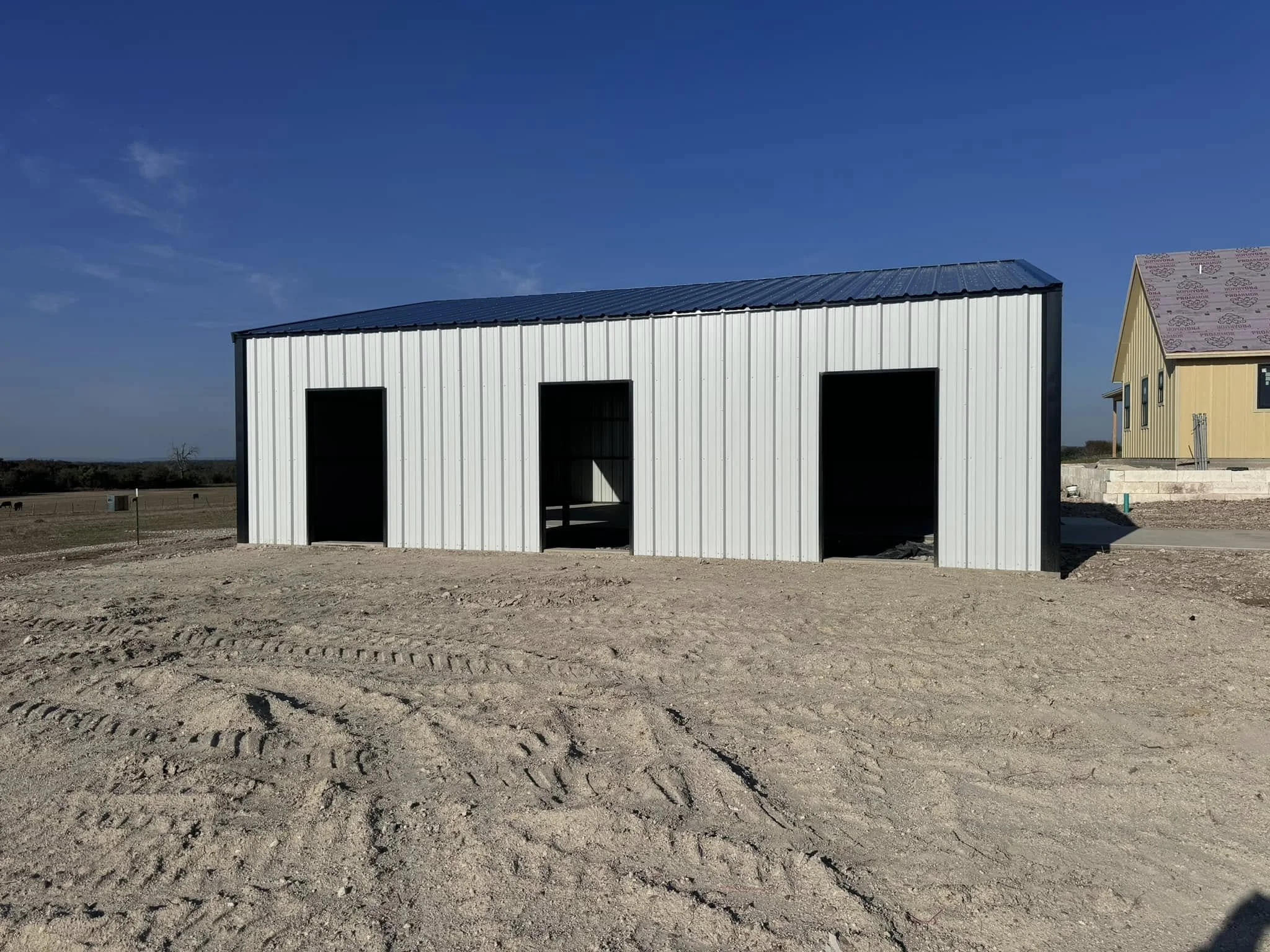 A partially constructed metal building with three large open doorways, situated on a sandy lot with visible tire tracks. A yellow house with a pink roof is in the background to the right.