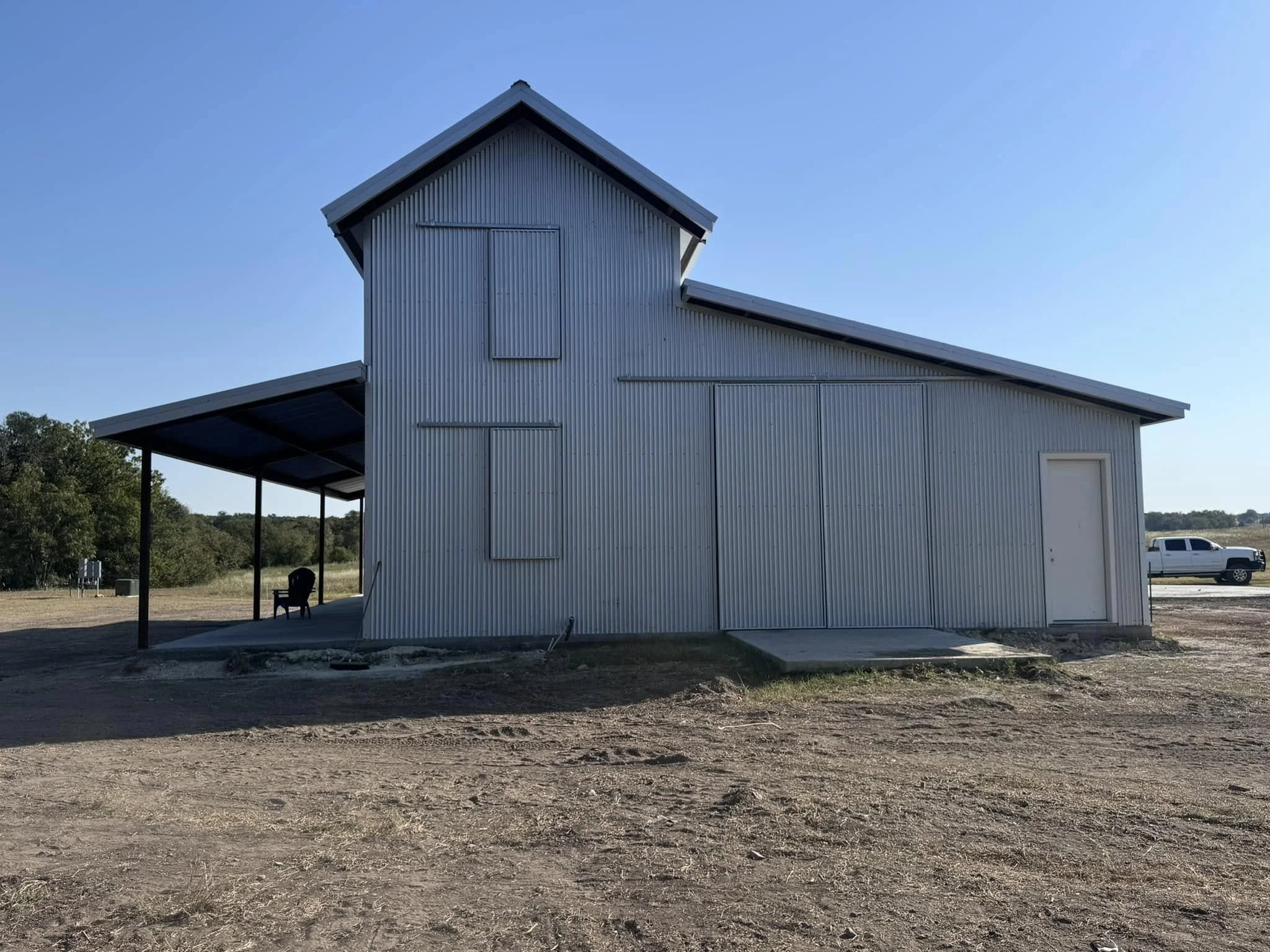 A large metal barn with a sloped roof and small upper window, surrounded by a dirt area, with trees in the background, and a white pickup truck parked nearby.