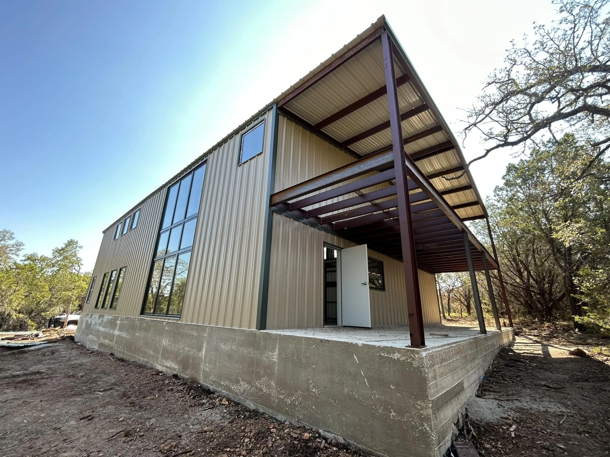 A modern metal building with a concrete foundation, large windows, and a raised porch with a metal roof supported by metal beams, surrounded by trees under a clear sky.
