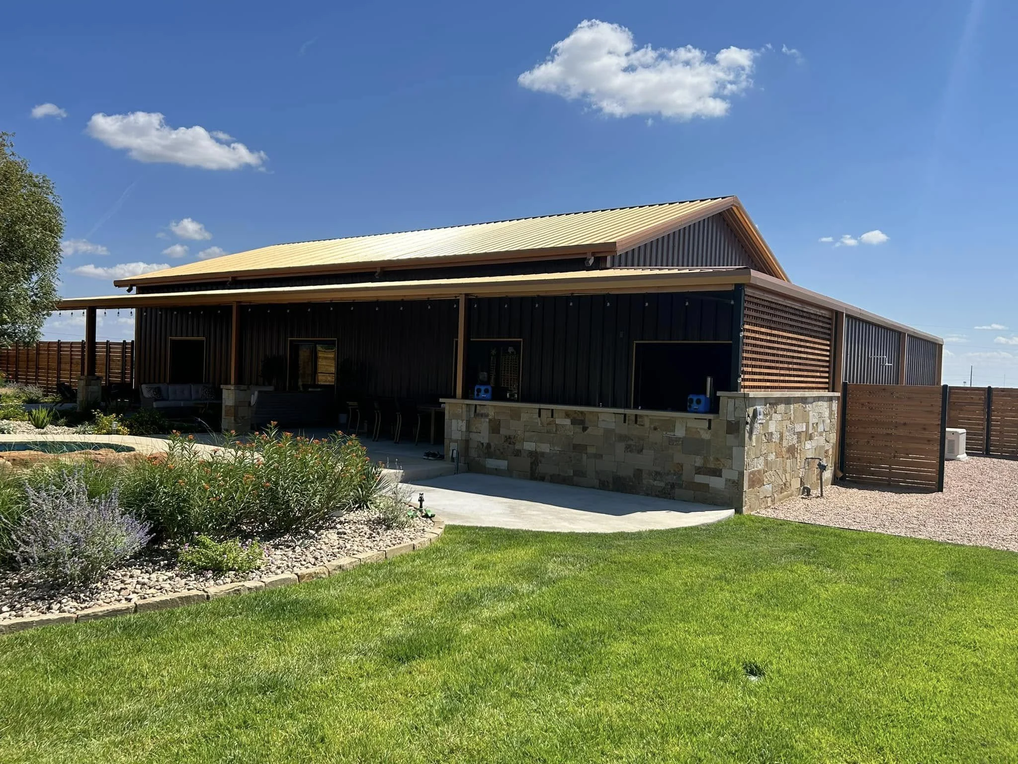 A modern house with a yellow metal roof, dark exterior walls, and a stone foundation, surrounded by a well-maintained lawn, garden plants, and a wooden privacy fence under a blue sky with a few scattered clouds.