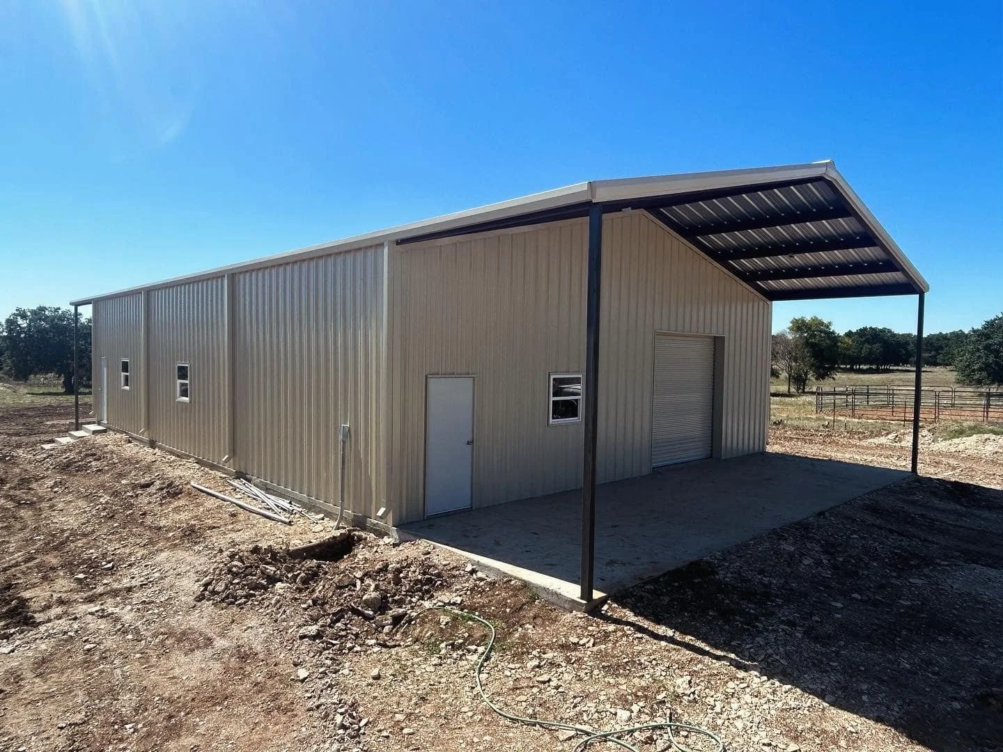 A beige metal shed with a sloped roof and a large roll-up door, situated on a dirt lot with a clear blue sky in the background.