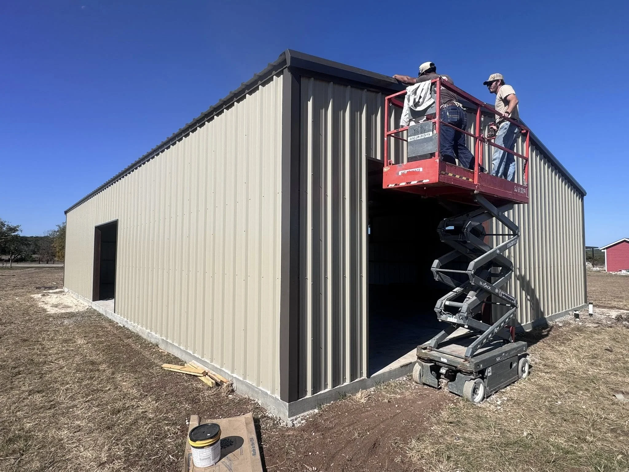 Two workers on a red scissor lift installing or repairing the roof of a beige metal storage building on a sunny day.
