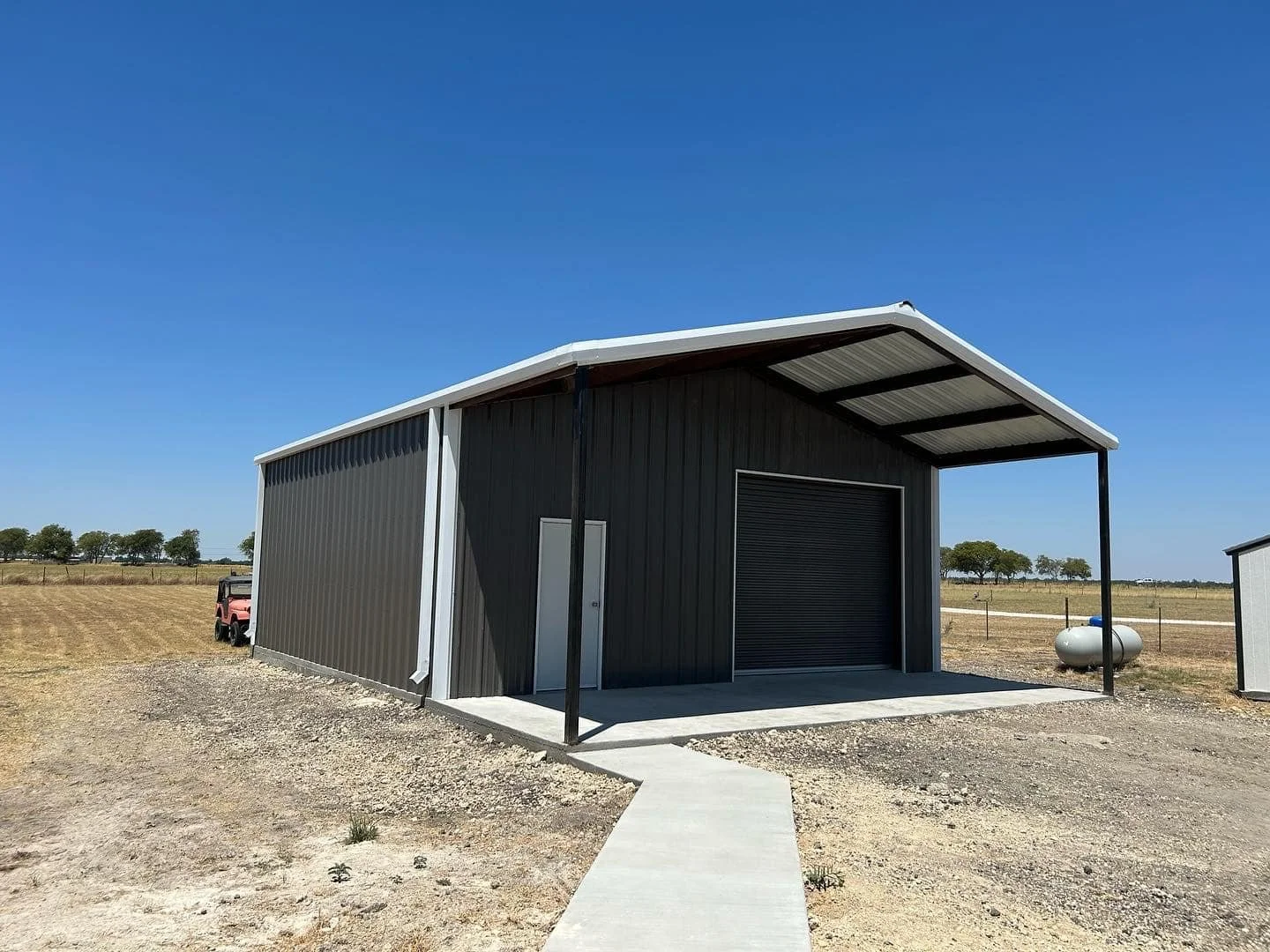 A black metal storage building with a roll-up door, a side door, and a pitched roof, located on a gravel lot in a rural area under a clear blue sky.