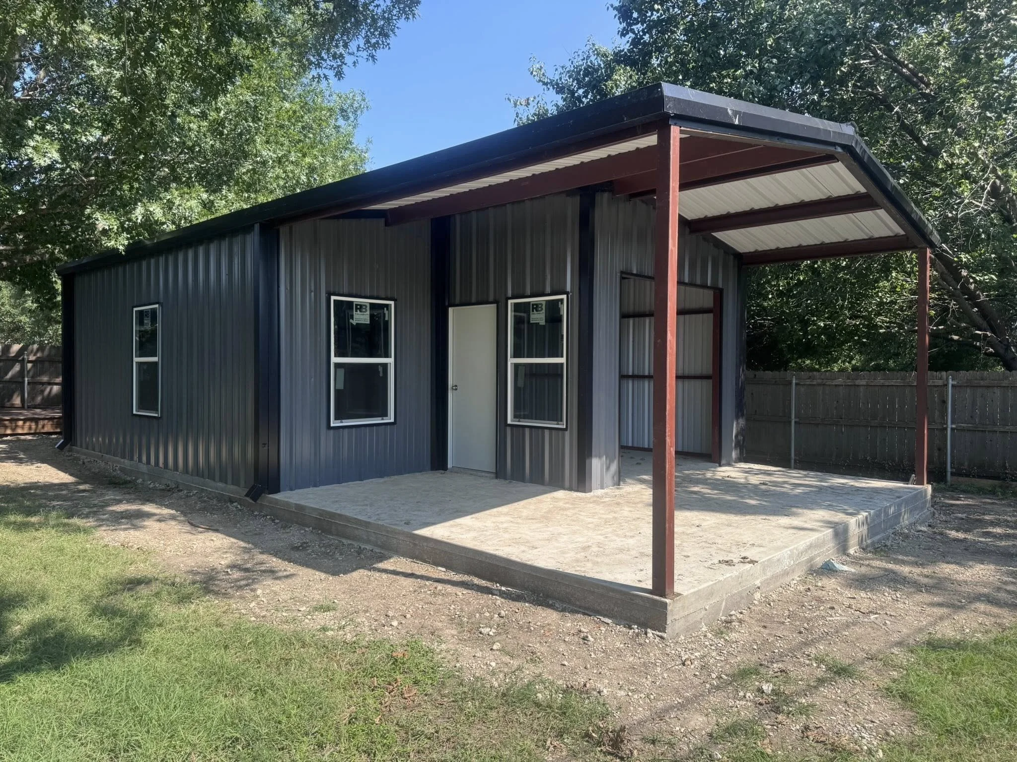 A new metal shed with a covered porch, surrounded by grass and trees in a backyard.