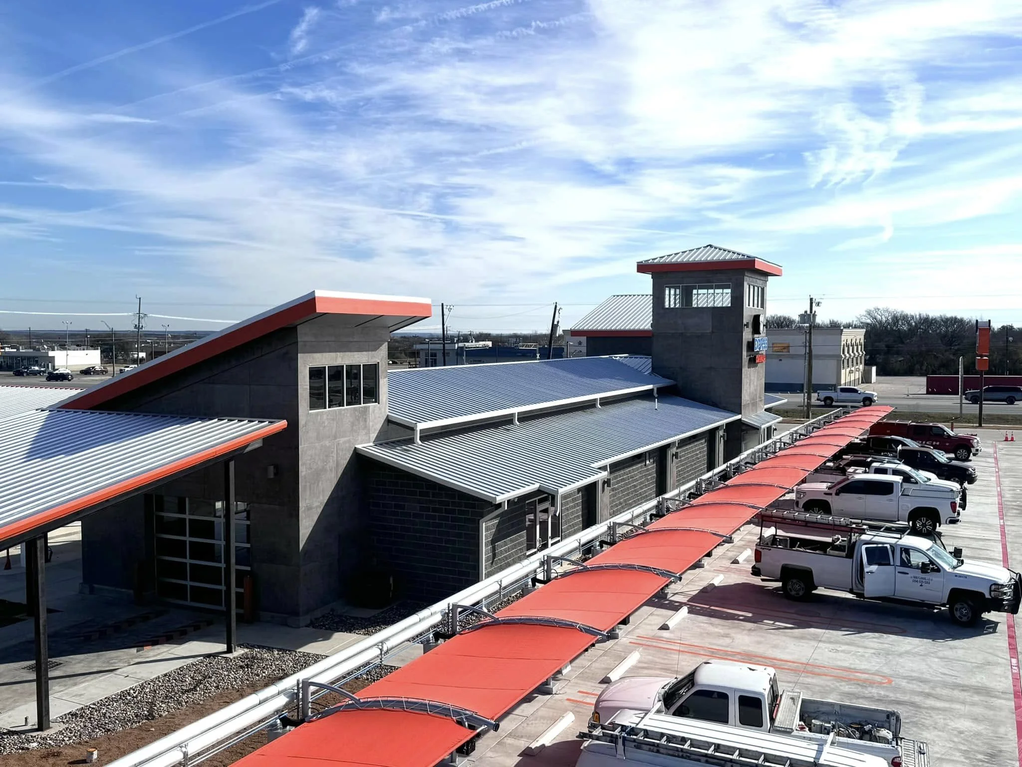 A modern commercial building with a metal roof and red accents, adjacent to a parking lot filled with pickup trucks and cars, under a partly cloudy sky.