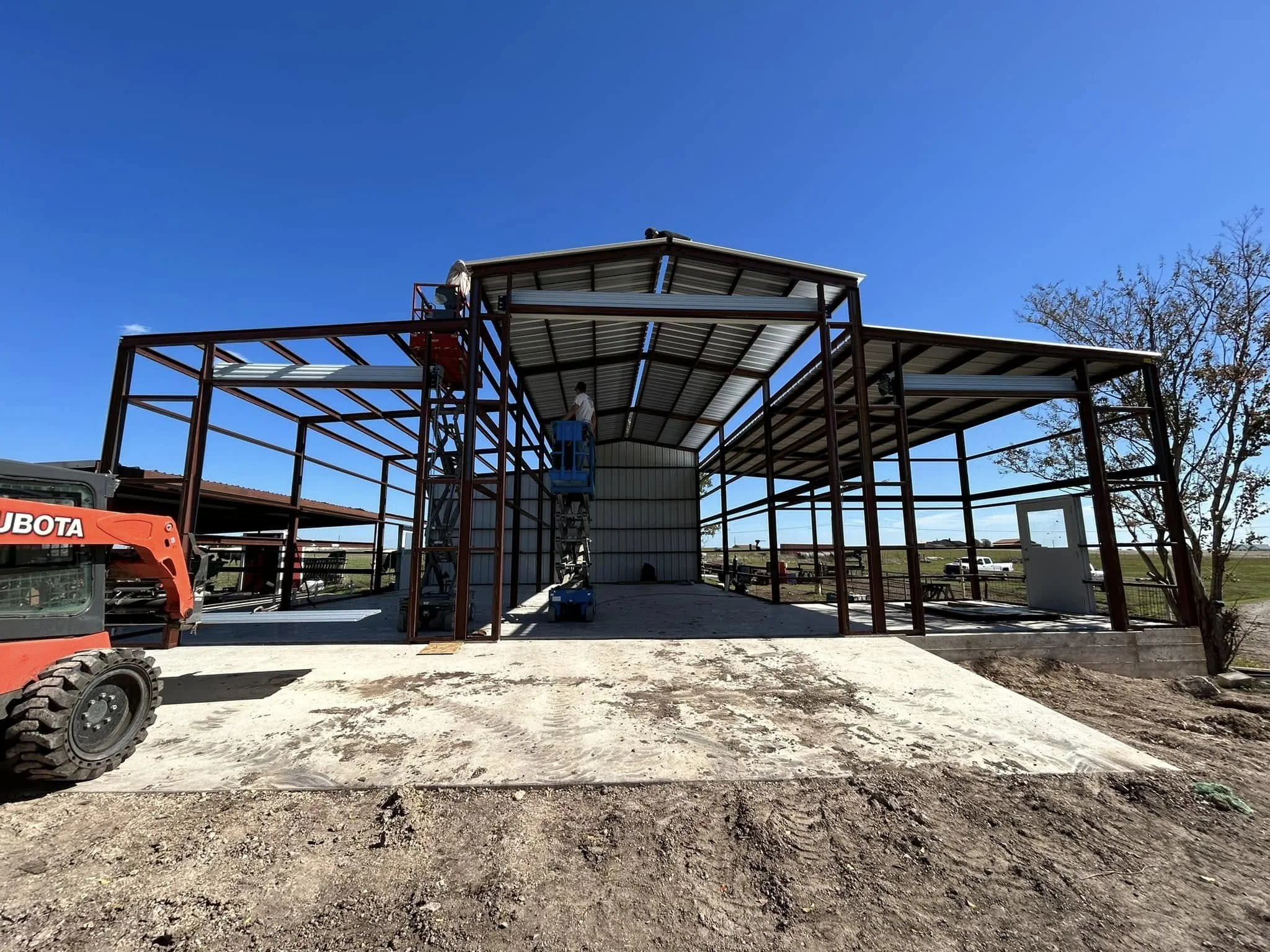 Construction site with a metal framework for a building, workers on lift equipment, a tree on the right, a blue sky background, and a small building in the distance.