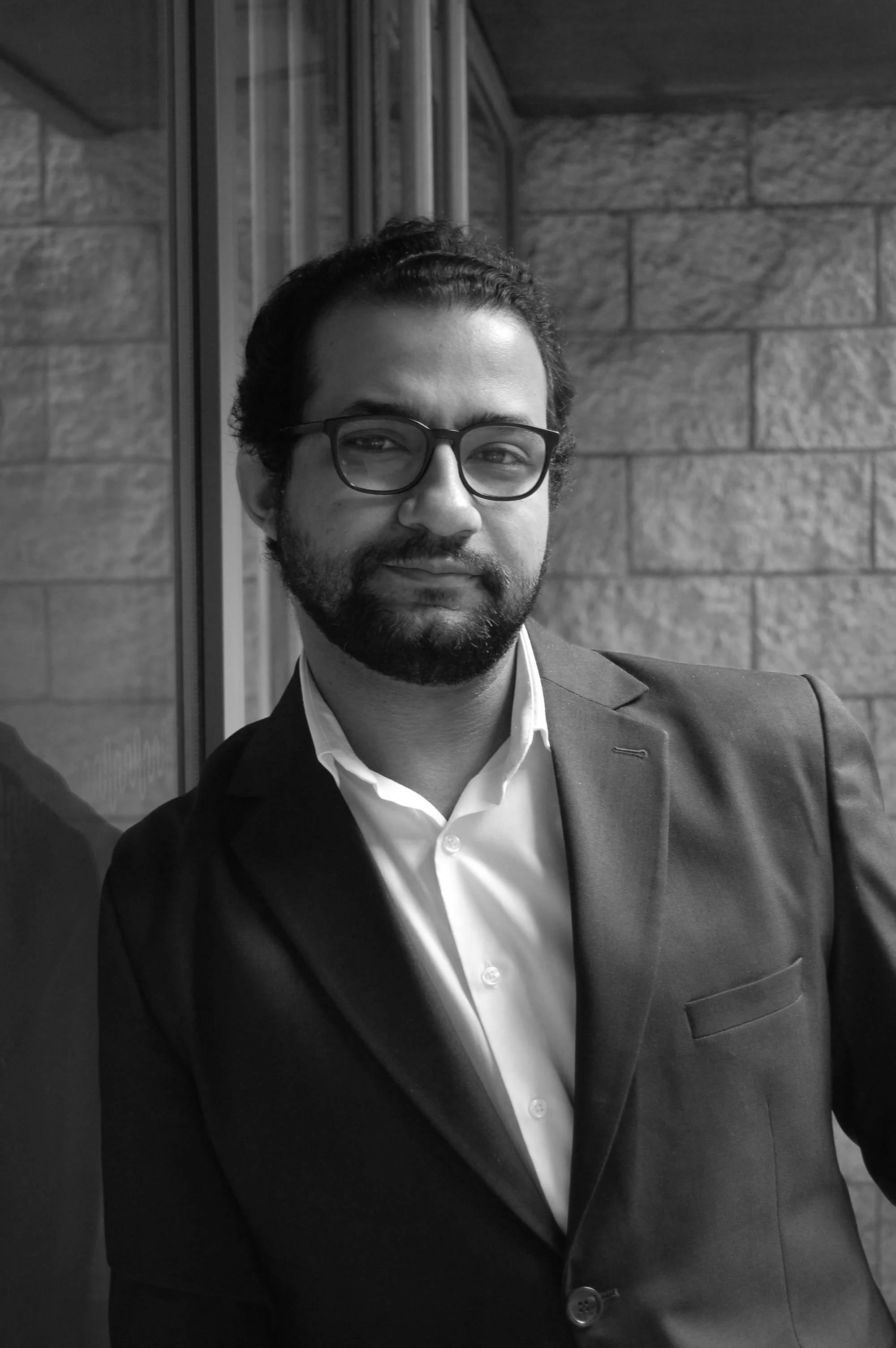 A man with glasses, a beard, and dark hair wearing a suit and white shirt, standing near a window with a brick wall background.