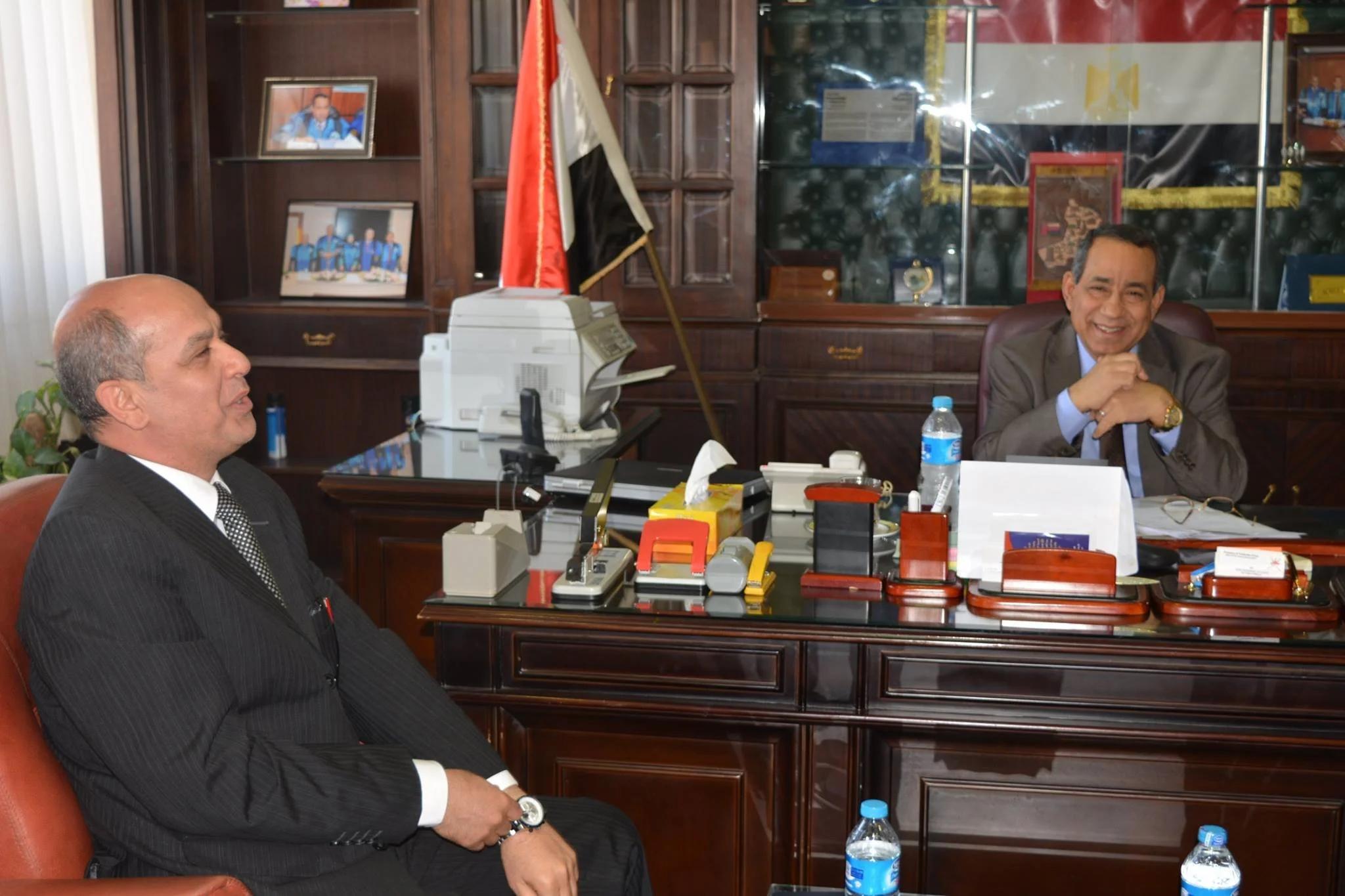 Two men in suits sitting in an office with dark wood panels and framed pictures on shelves; one is smiling with his hand on his chin, the other is sitting with closed eyes and hands clasped in his lap; on the desk are office supplies and water bottles, and a flag is behind one man.