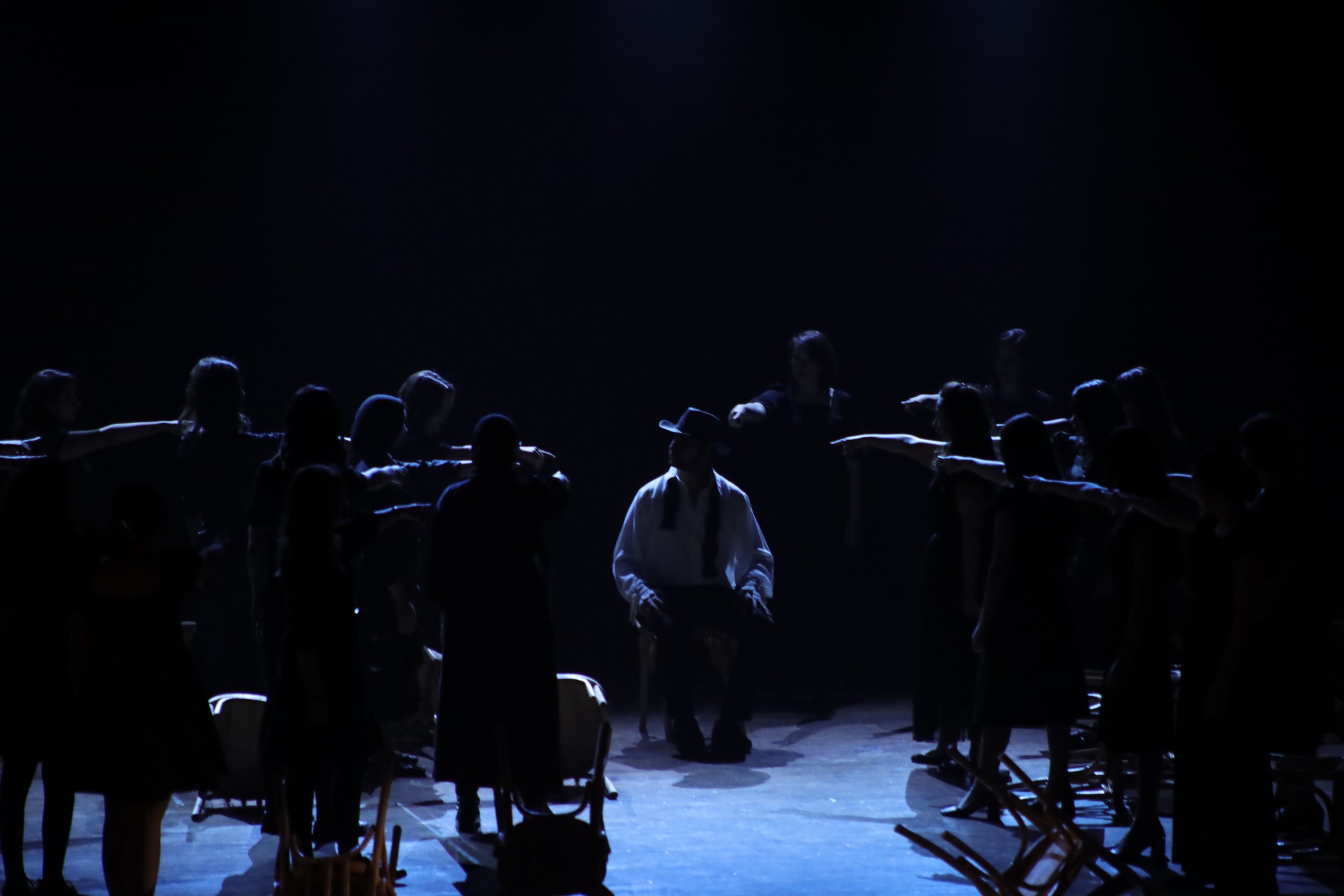 Stage performance with silhouetted performers arranged in a semi-circle, illuminated by minimal lighting, creating a dramatic and mysterious atmosphere.
