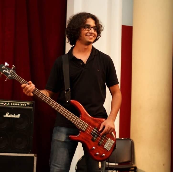 Young man with curly hair, glasses, wearing a black polo shirt and jeans, holding a red bass guitar, standing next to a Marshall amplifier in a room with red and beige curtains.