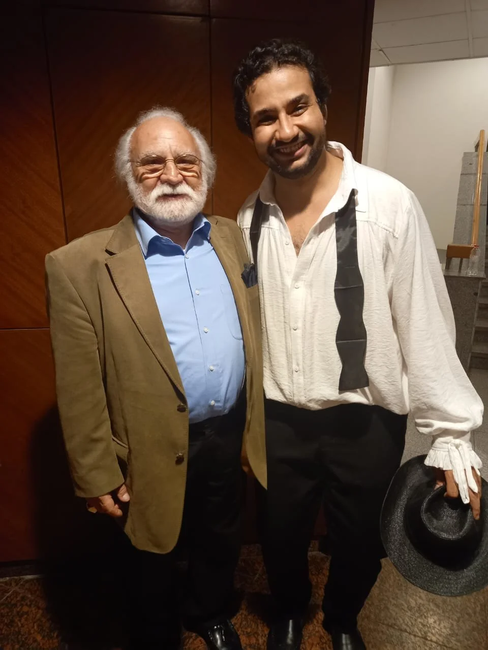 Two men smiling, standing close together indoors, one with white hair and beard wearing a tan blazer and blue shirt, the other with dark curly hair and beard wearing a white shirt with a black ribbon detail, holding a black hat and a pair of glasses.