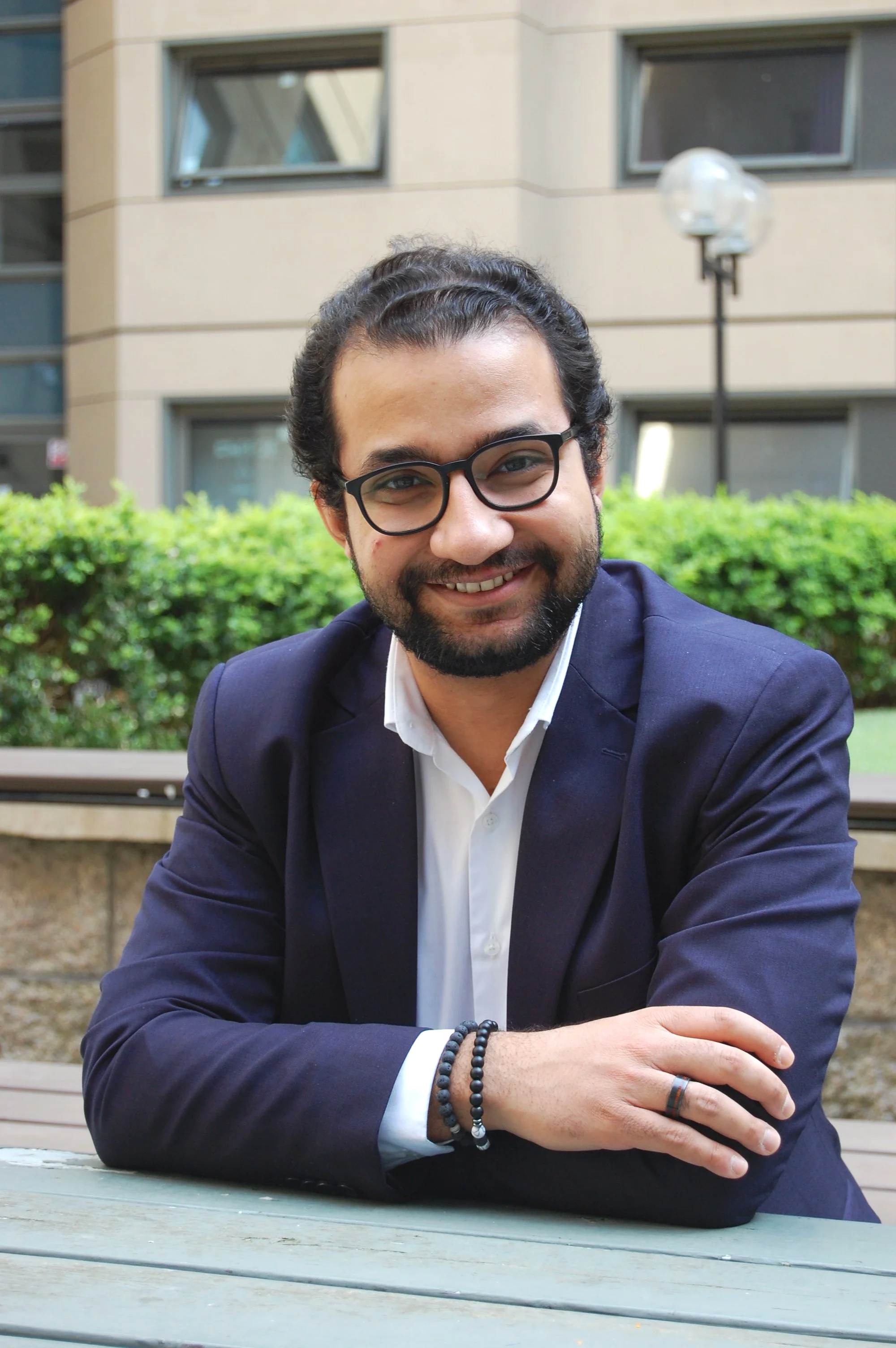 A man with glasses, a beard, and dark curly hair sitting outdoors at a table. He is wearing a dark blazer, a white shirt, and beaded bracelets. The background shows green bushes and a building with rectangular windows and a streetlamp.