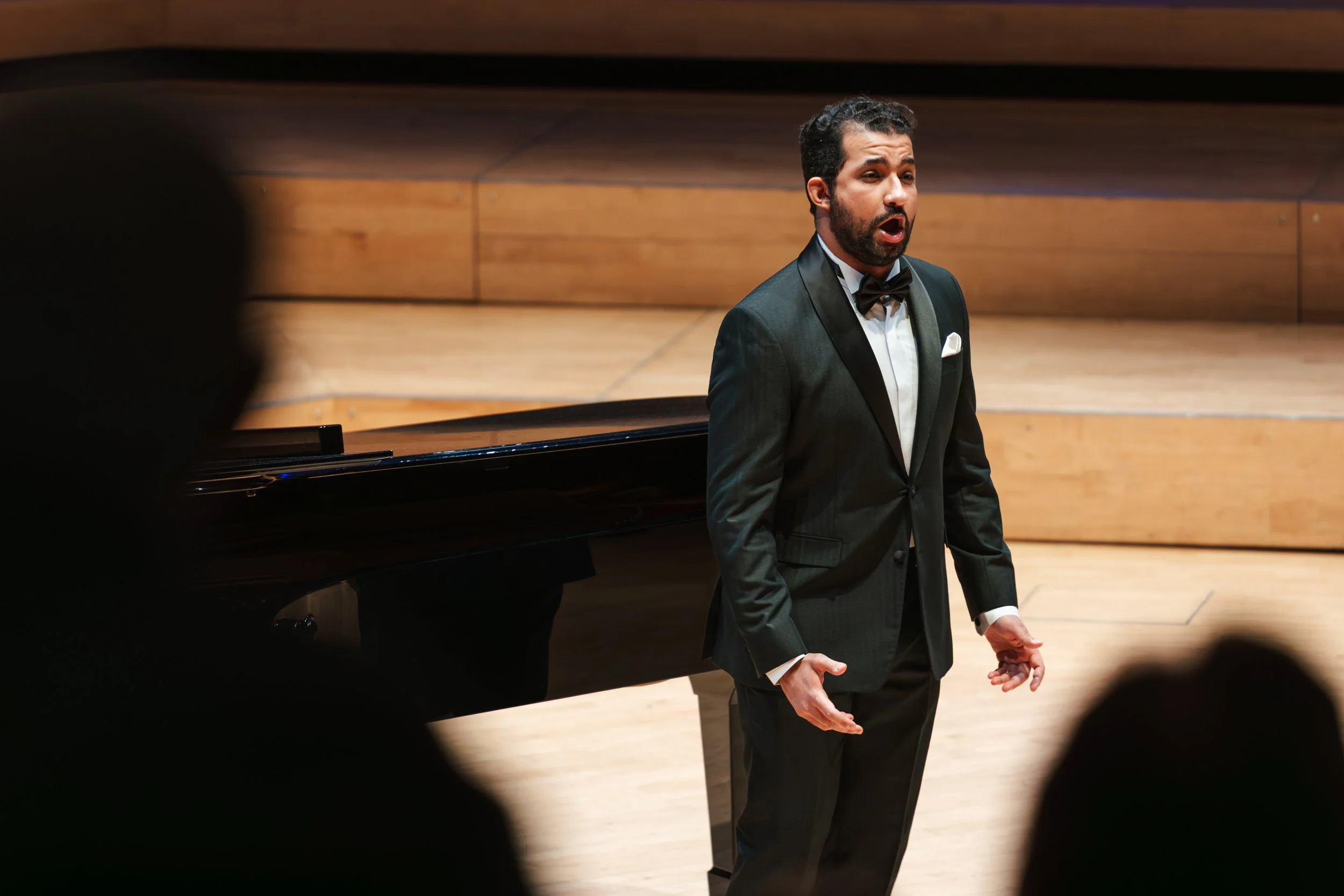 A man in a tuxedo singing passionately in a concert hall with a grand piano nearby.