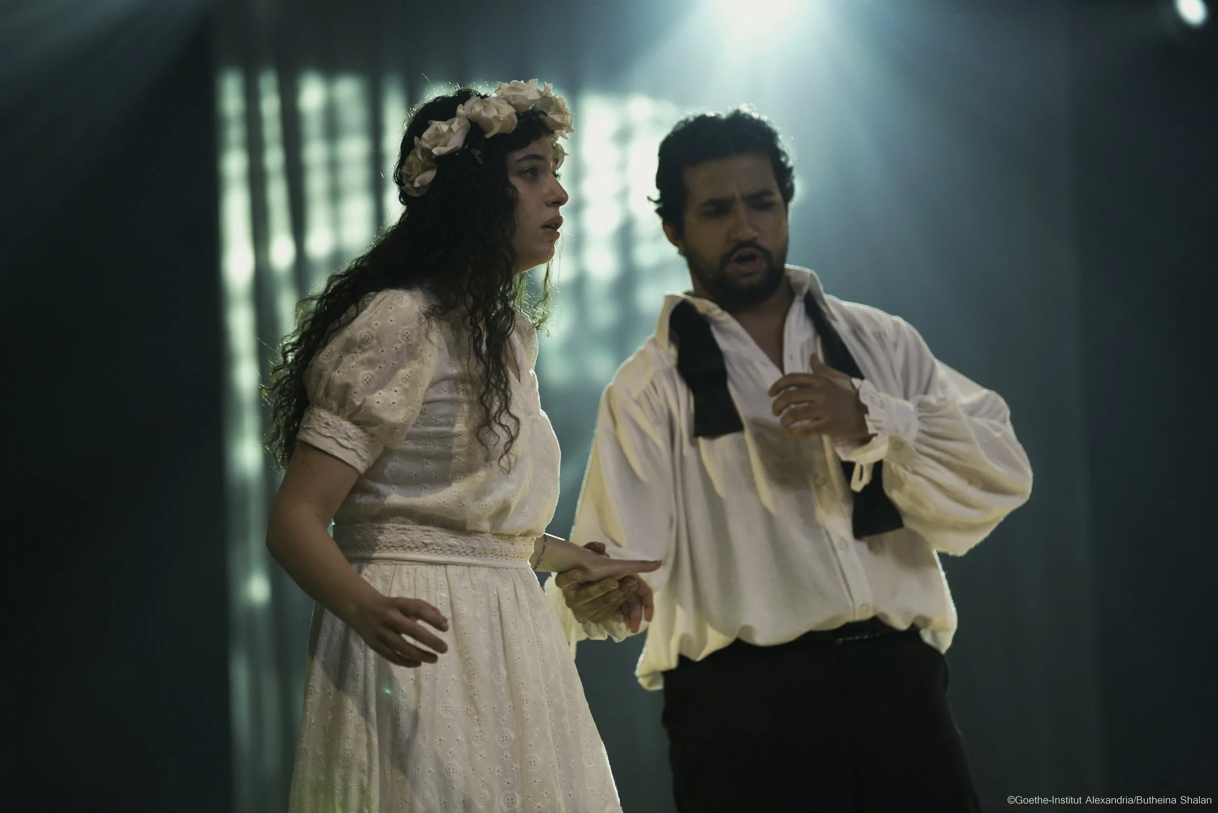 A woman wearing a white dress and a flower crown is holding hands with a man wearing a white shirt and black pants. They appear to be in an emotional or serious moment, with soft lighting and a blurred background.