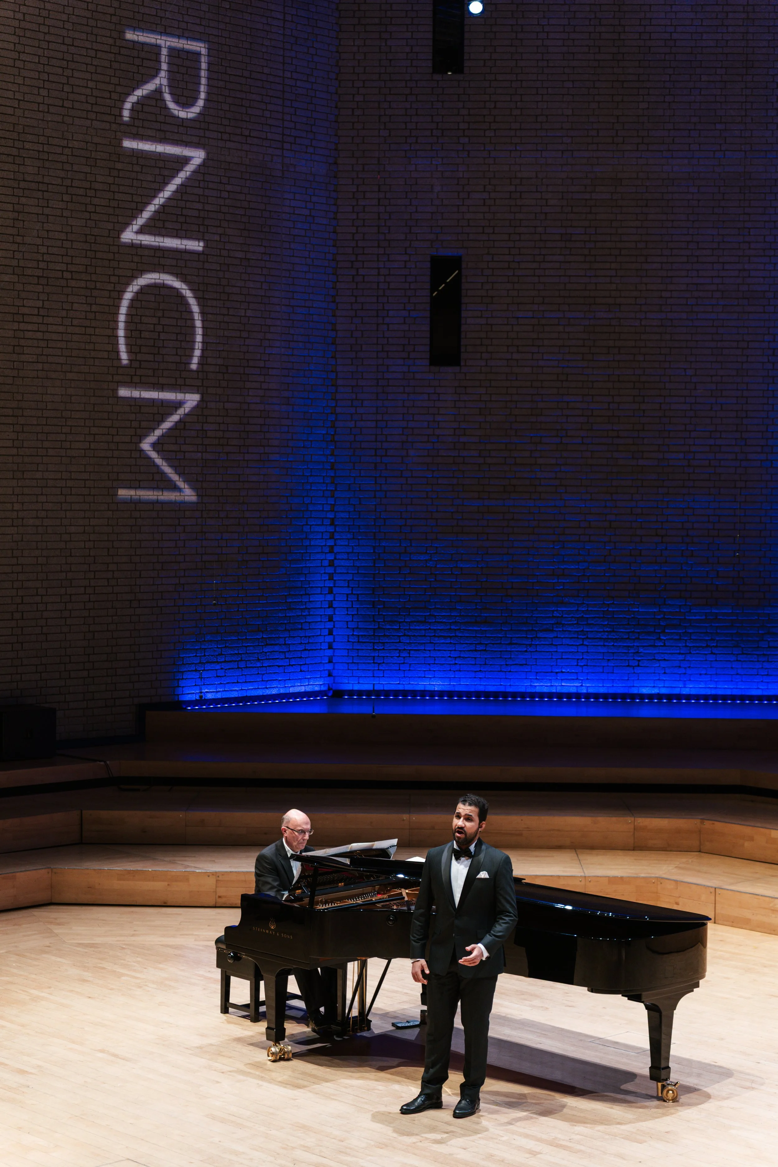 A classical music performance on stage with a male singer in a tuxedo, a pianist at a grand piano, and a wooden stage with a brick wall backdrop illuminated by blue lighting. The word '2023' is projected on the wall.
