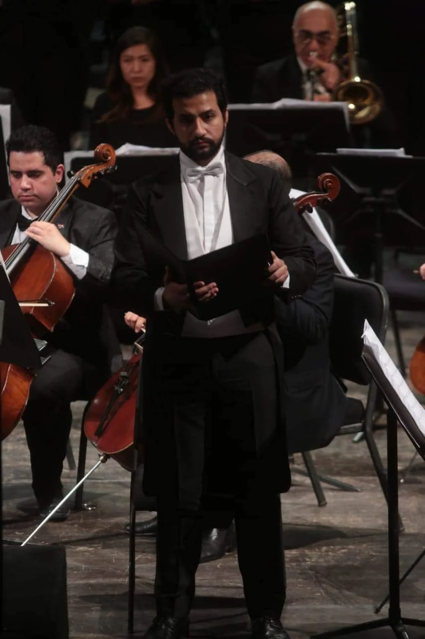 Classical orchestra conductor in black tuxedo with white shirt and bow tie, holding a music folder, surrounded by seated musicians playing string instruments and a musician with a trumpet in the background.