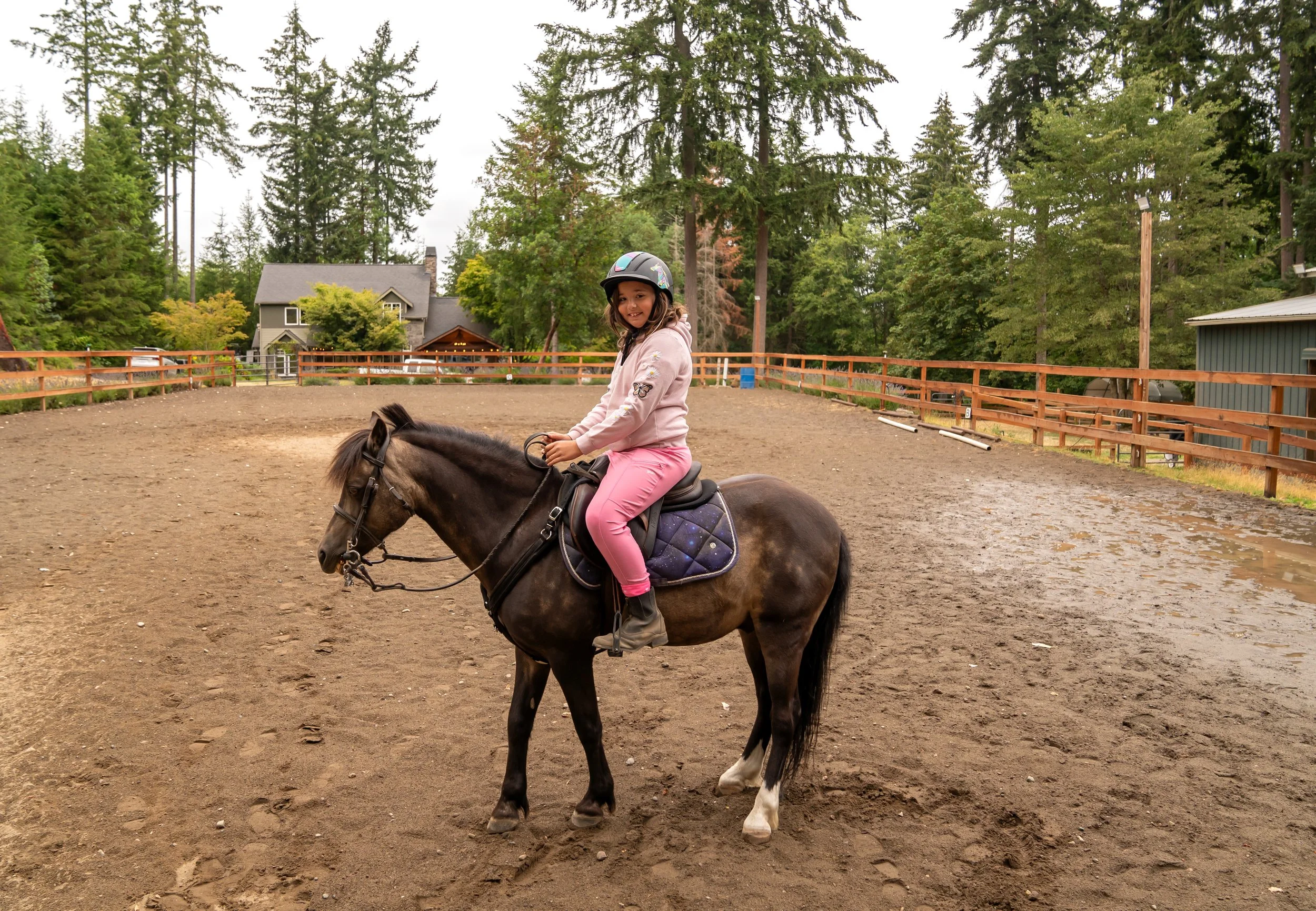A young girl riding a brown horse in an outdoor riding arena, surrounded by tall trees and houses in the background, on a cloudy day.