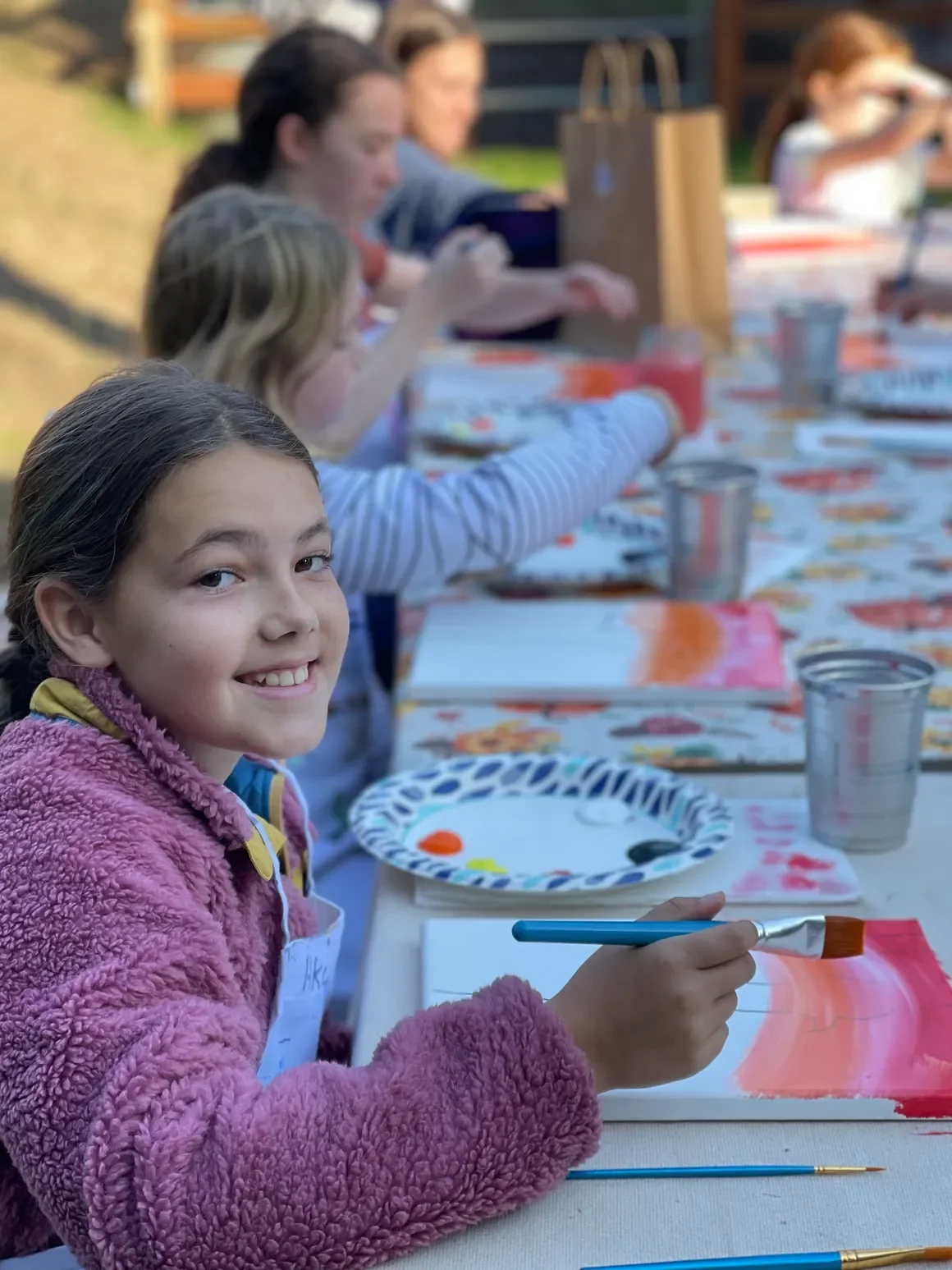 A young girl with dark hair in a ponytail, wearing a pink fuzzy jacket, smiling at the camera while holding a paintbrush and painting on a white canvas. She is sitting at a table outdoors with other children, and the table has art supplies, paper plates with paint, and cups of water.