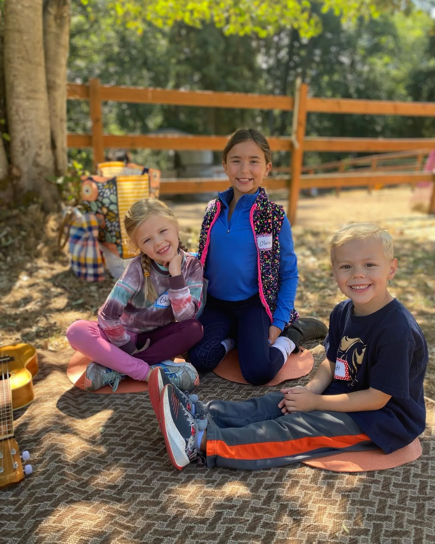 Three children sitting on mats outdoors, smiling. One girl has braided hair, and a boy has blond hair. They are in a sunny area with trees and a wooden fence in the background.