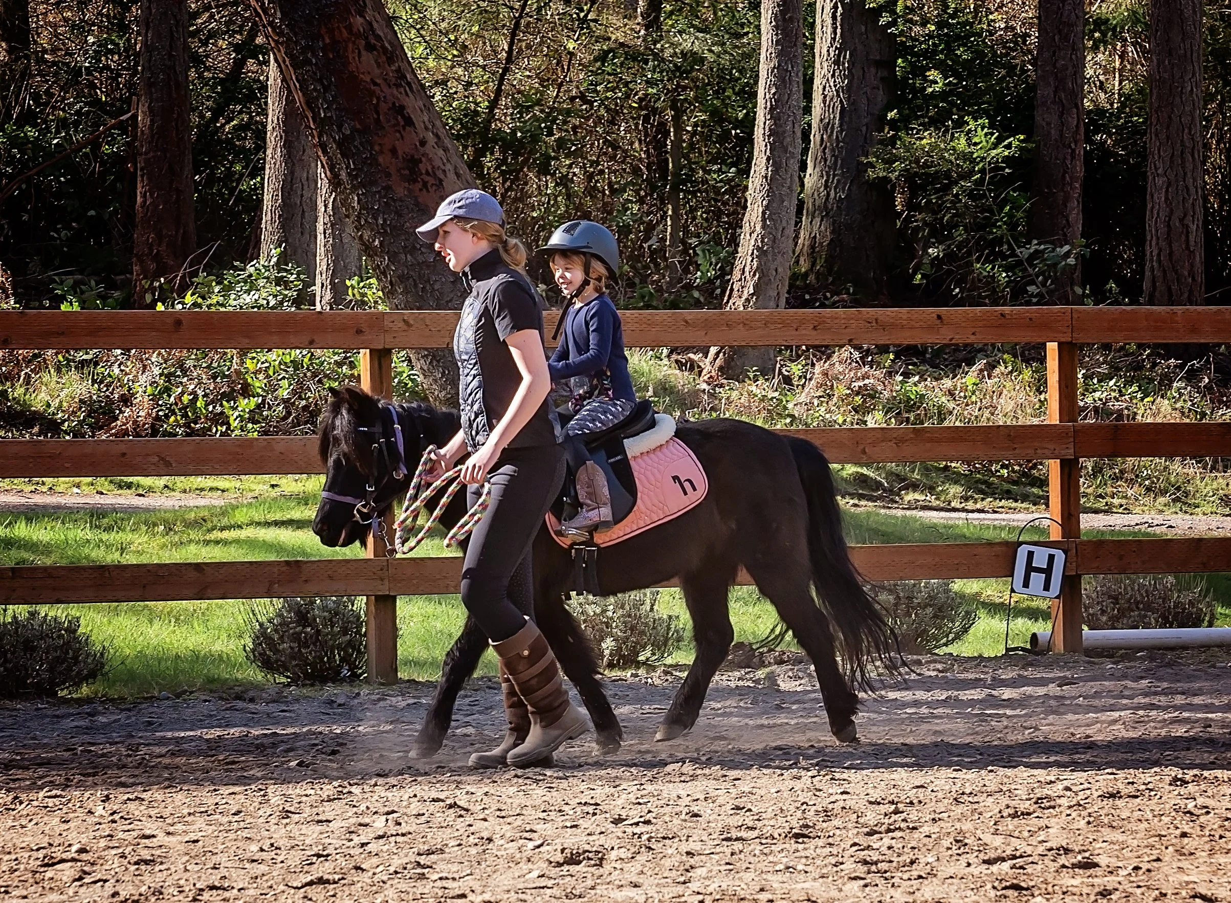 Two young girls riding a small horse in an outdoor riding arena surrounded by a wooden fence, trees, and grass.