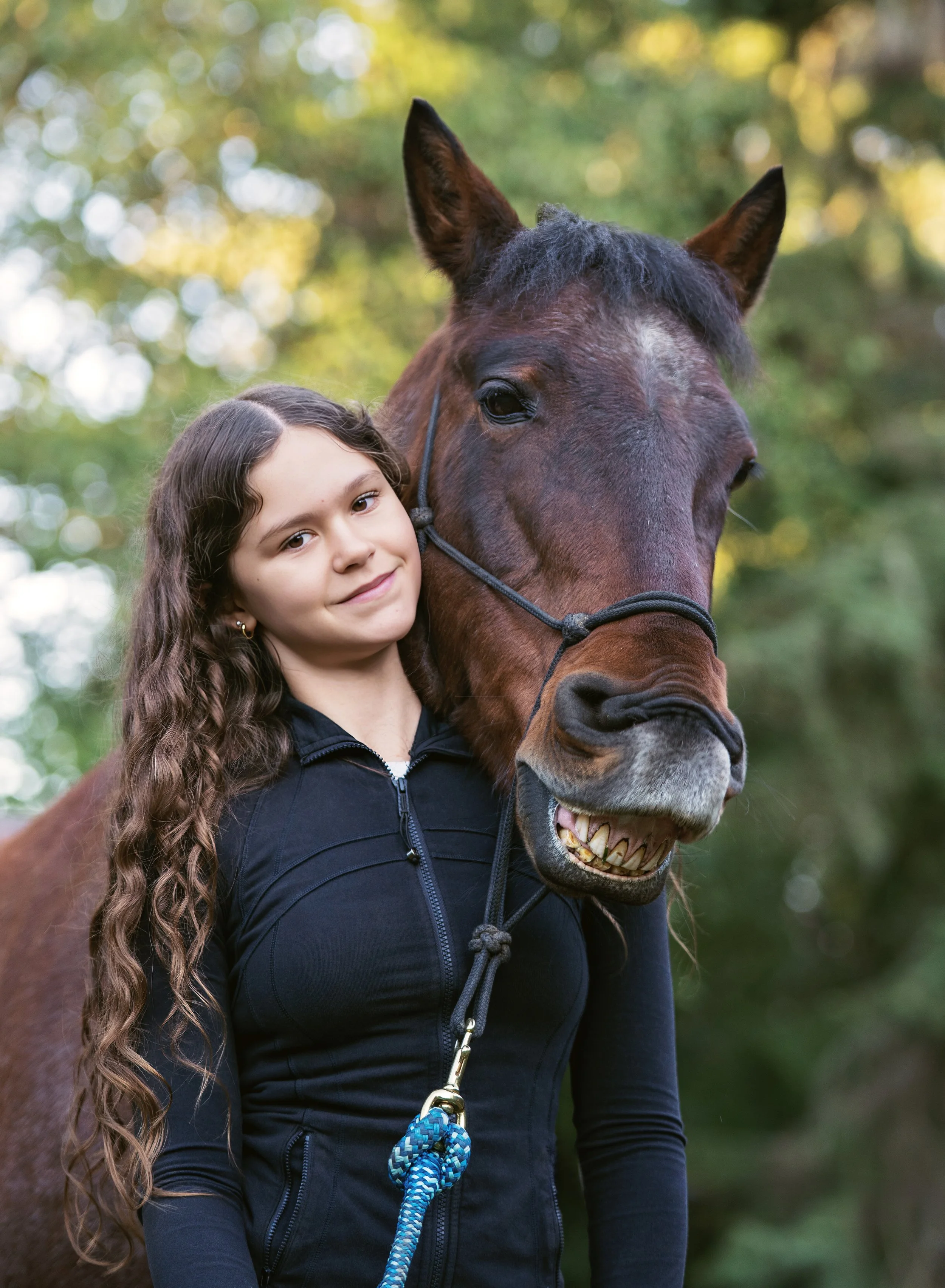 A girl with long, curly brown hair standing next to a brown horse with a smiling expression in a wooded outdoor setting.