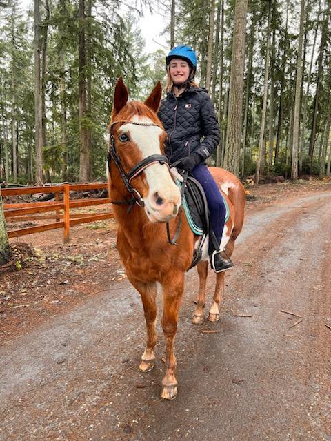 A smiling woman wearing a blue helmet and black jacket riding a chestnut horse with a white blaze down a dirt trail in a wooded area.