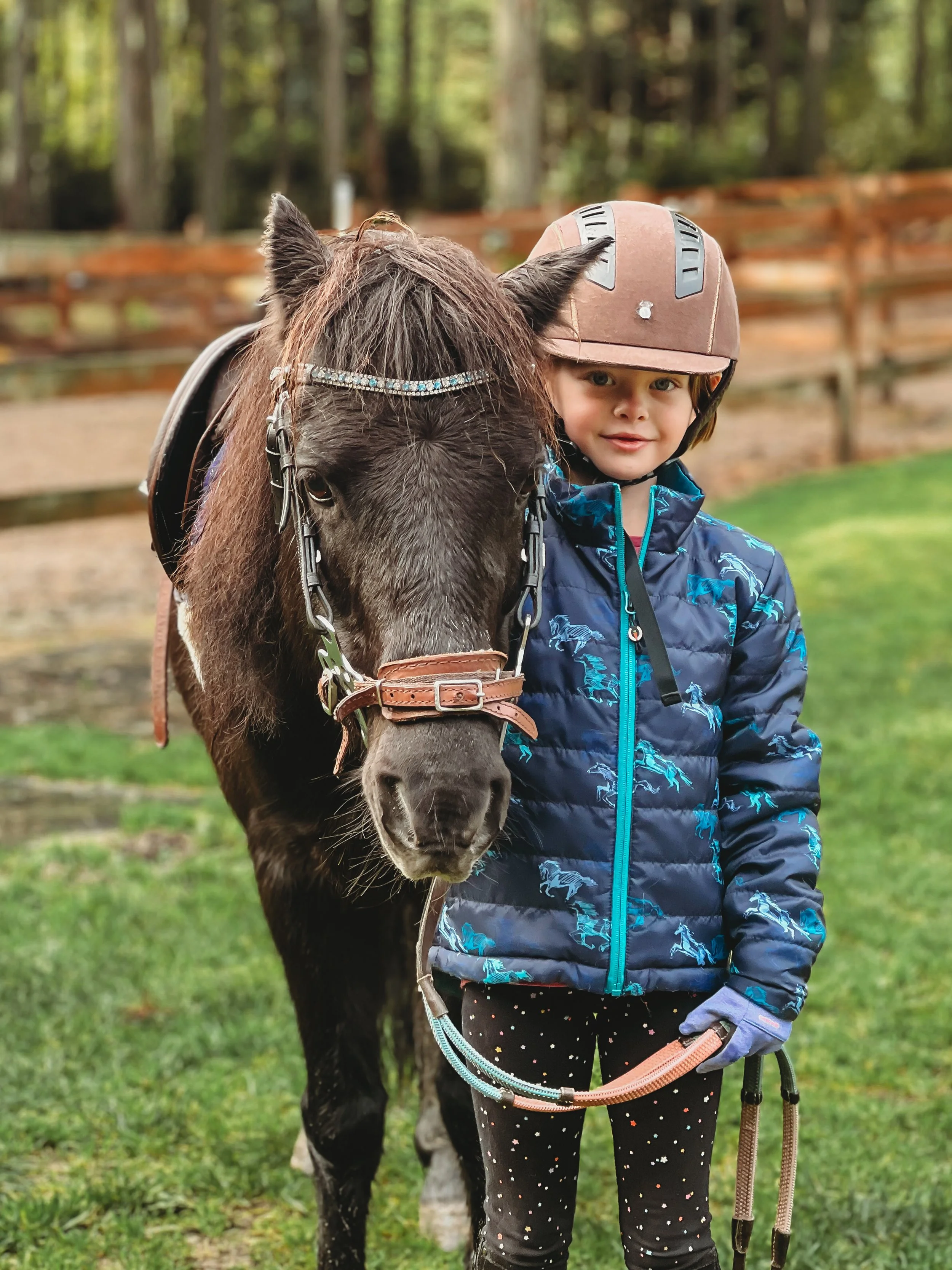 A young girl wearing a helmet and a blue jacket standing next to a black pony with a bridle and decorated with a sparkly browband.